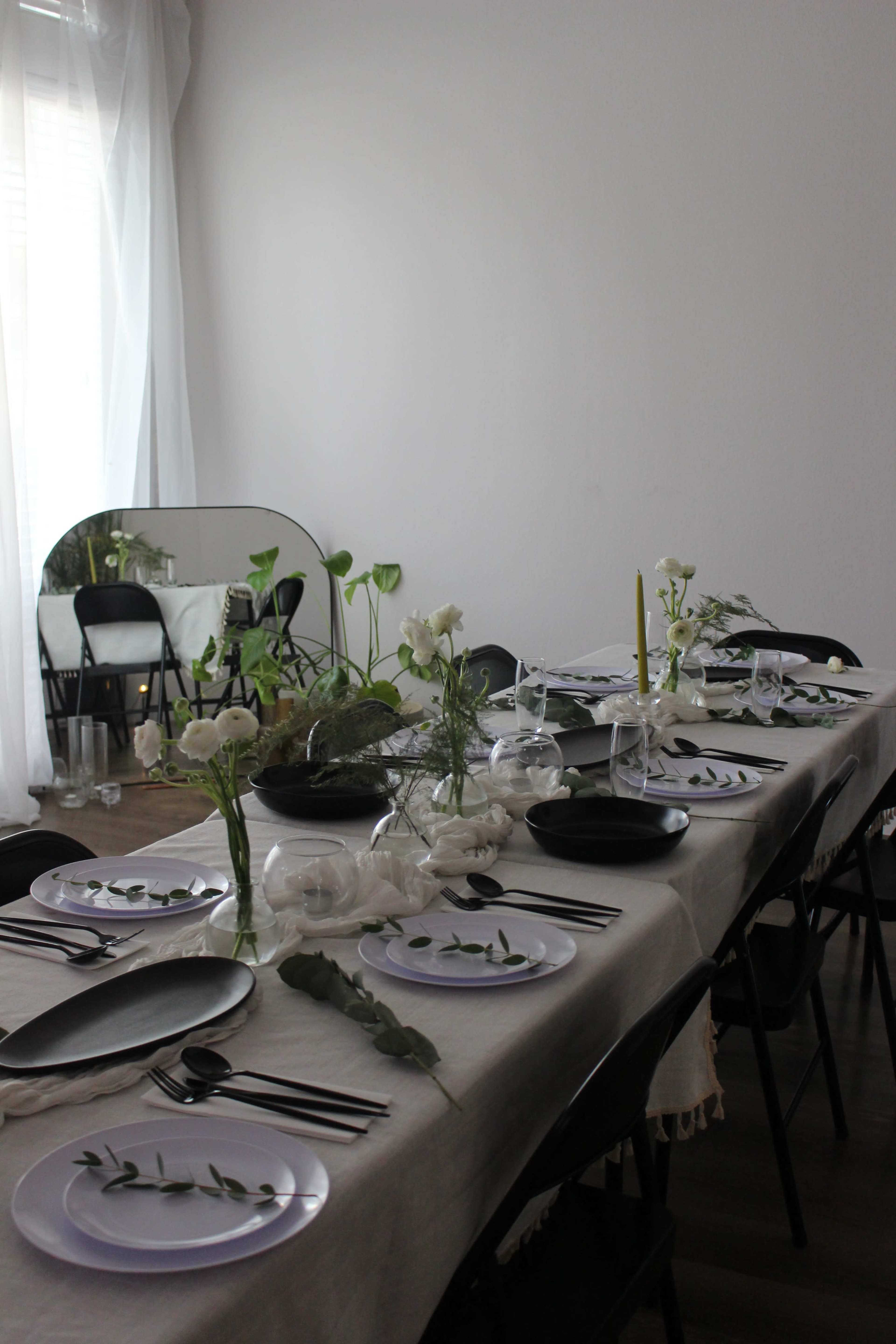 A long dining table is set with white and black plates, cutlery, and elegant floral arrangements.