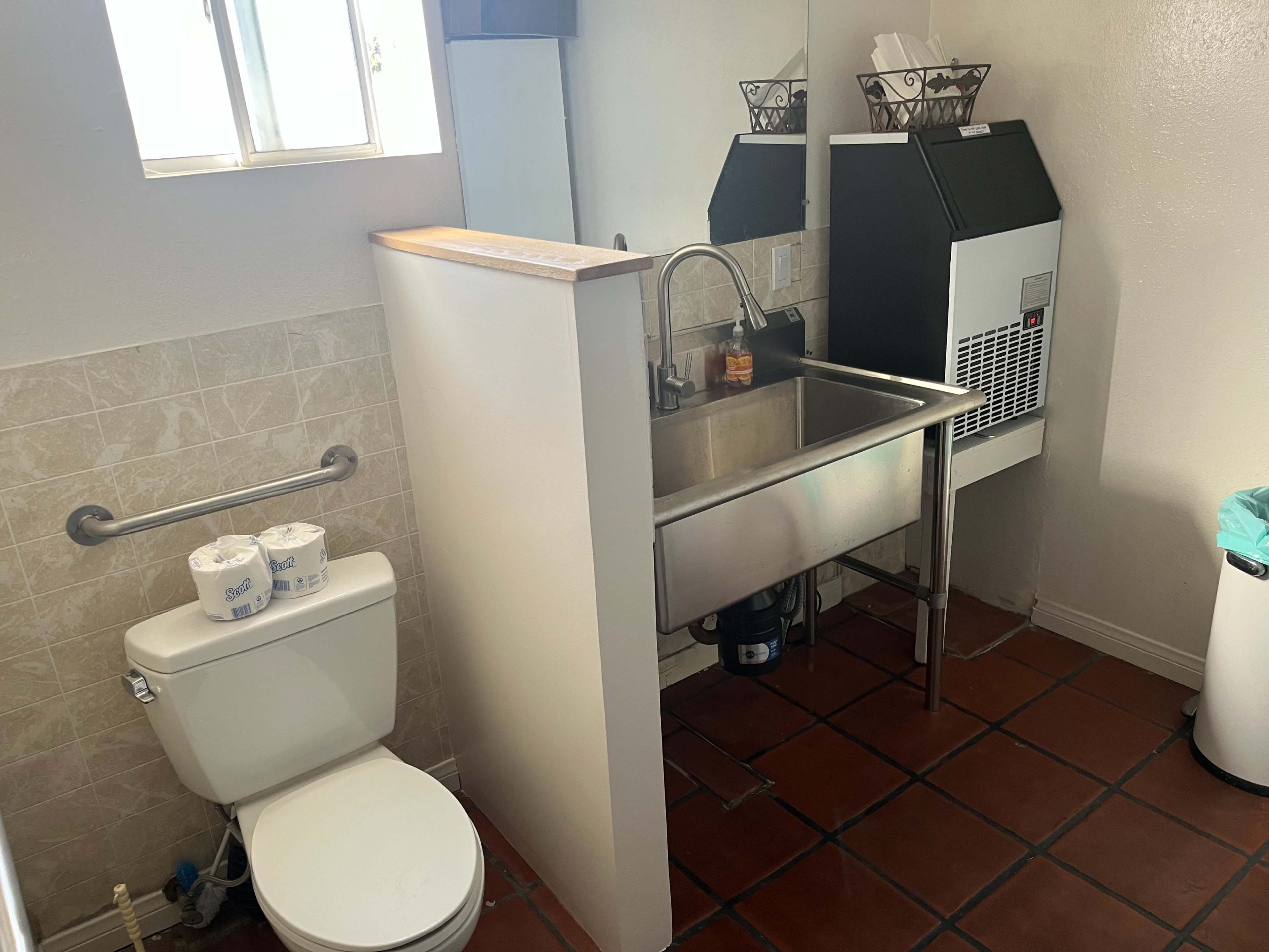 A small bathroom with a toilet, a stainless steel sink, and an ice maker, along with a trash can in the corner.