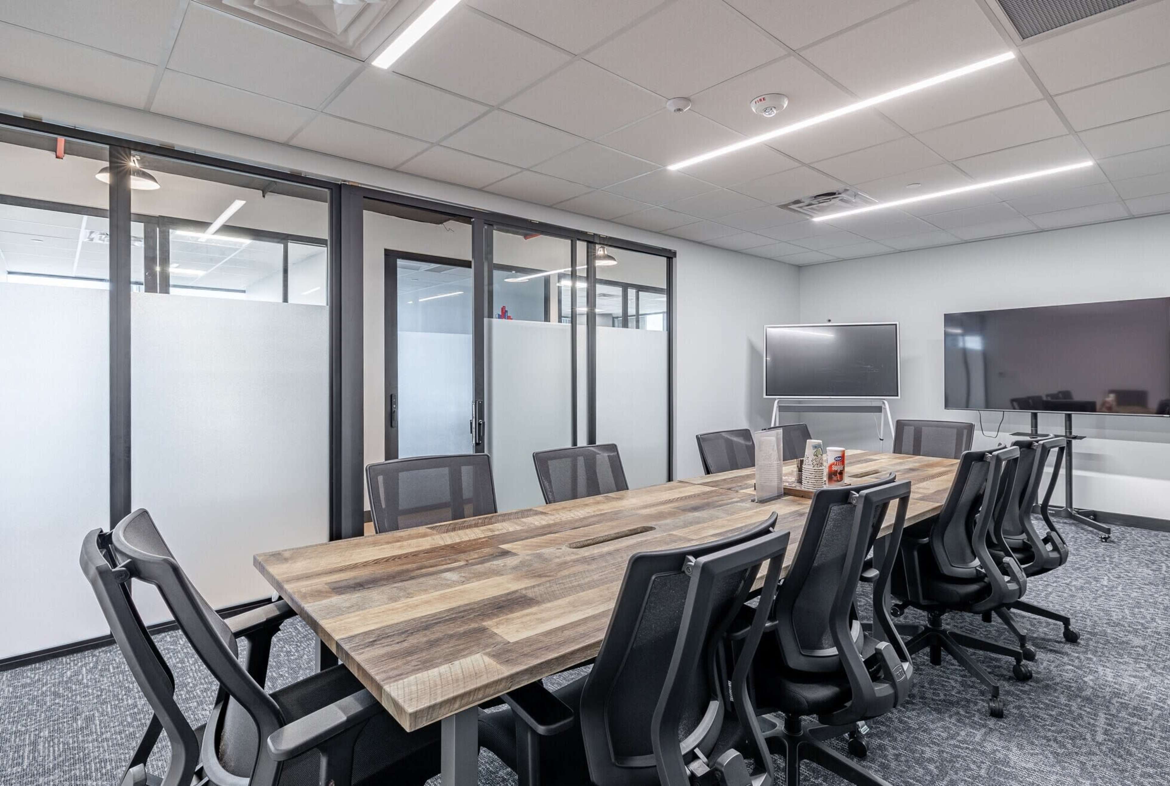 A modern conference room features a long wooden table surrounded by black office chairs, with glass partitions and large screens on the walls.