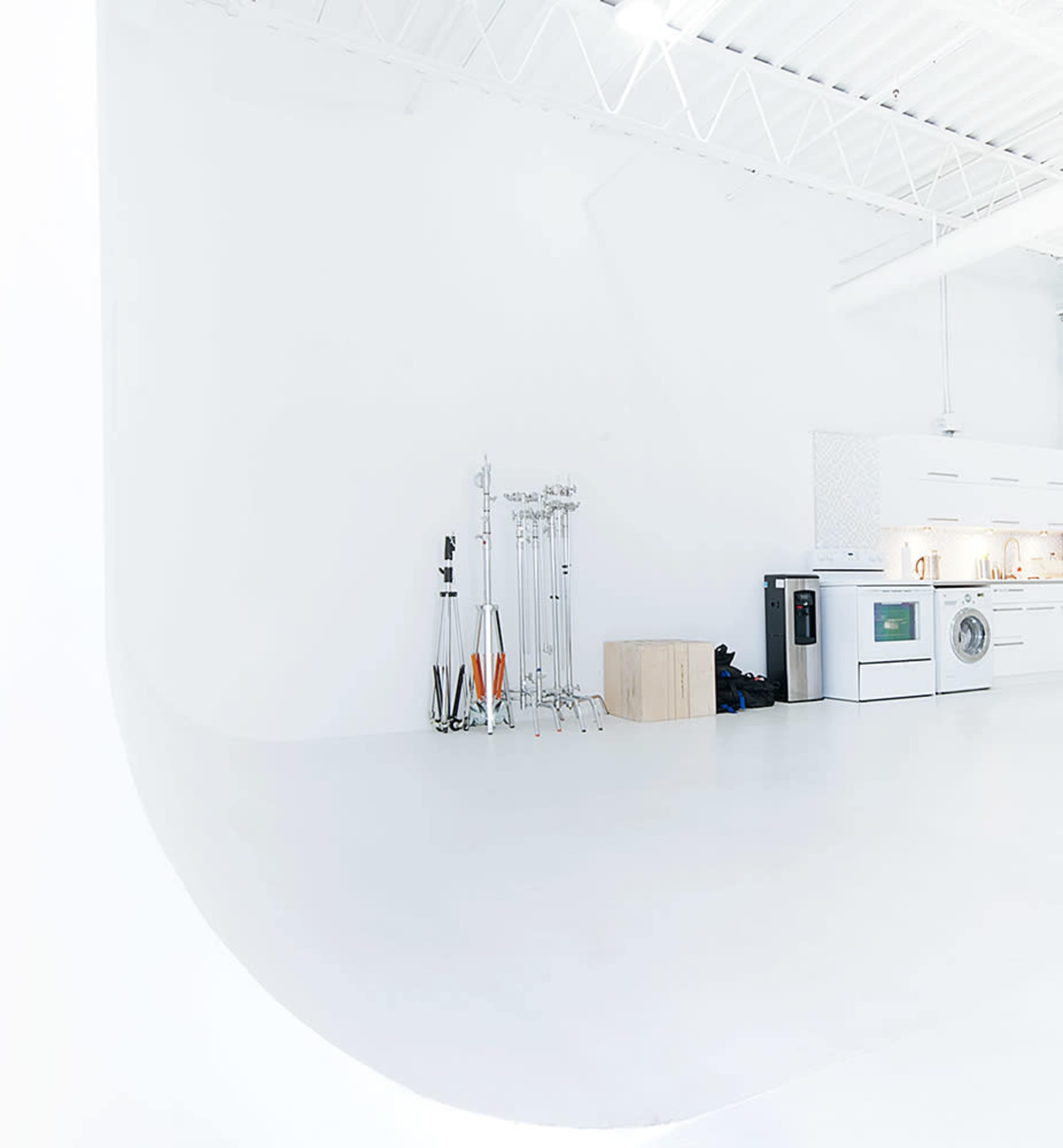 The image shows a minimalist studio space with a white curved wall, a small kitchen area, and equipment stands neatly arranged in a corner.