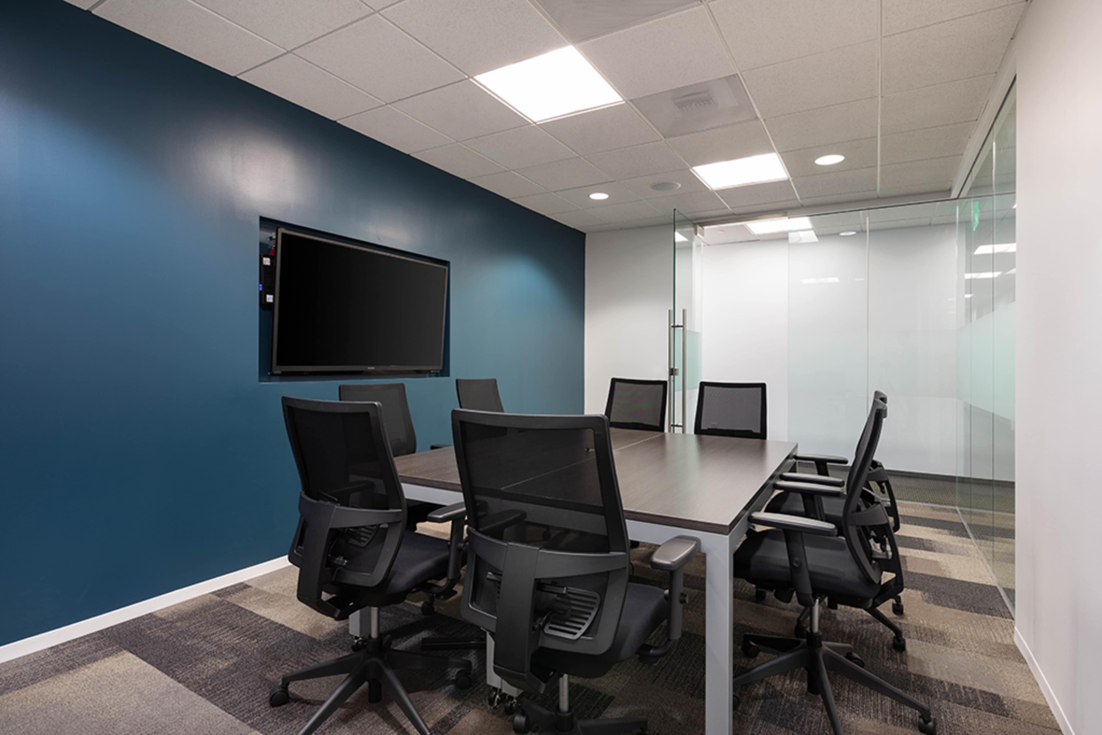 A modern conference room features a long table surrounded by black chairs and a large wall-mounted screen on a dark blue accent wall.