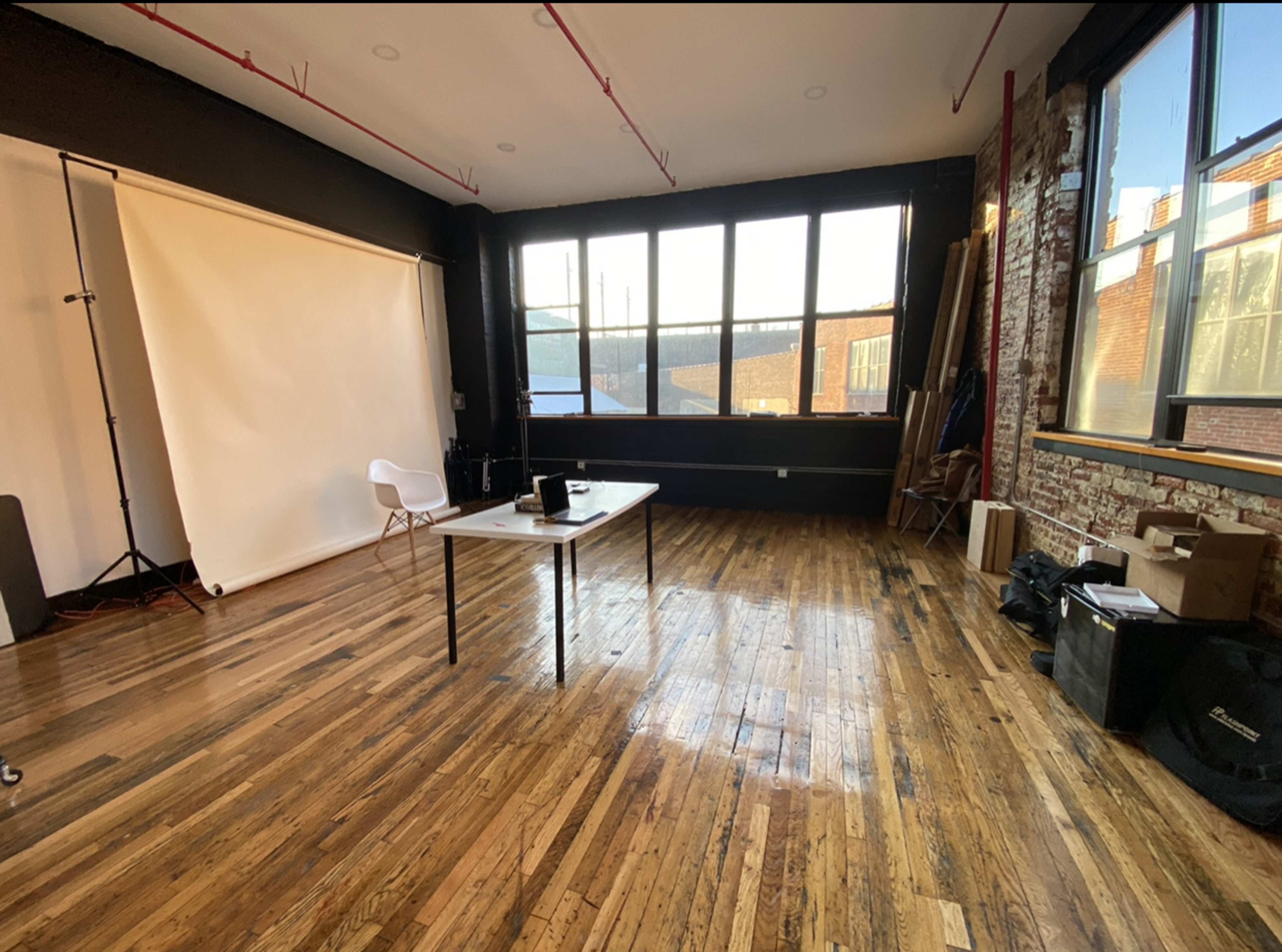 The image shows an empty photography studio with wooden floors, a table in the center, and large windows allowing natural light to enter.