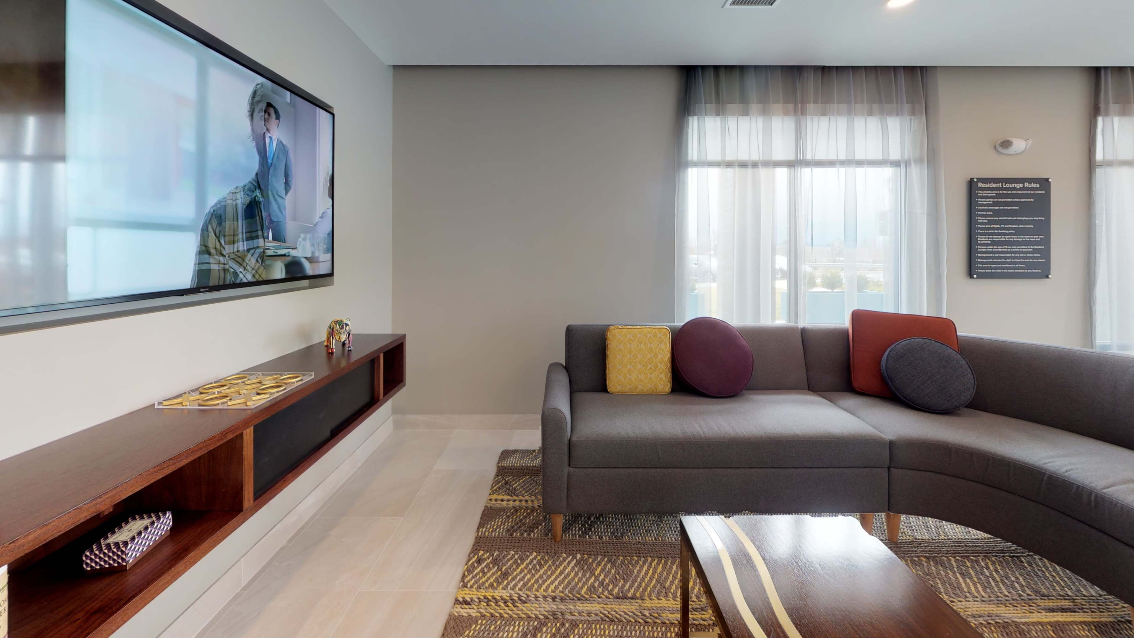 A modern living area features a sectional gray sofa, a wall-mounted television, and a wooden coffee table in front of a large window with sheer curtains.