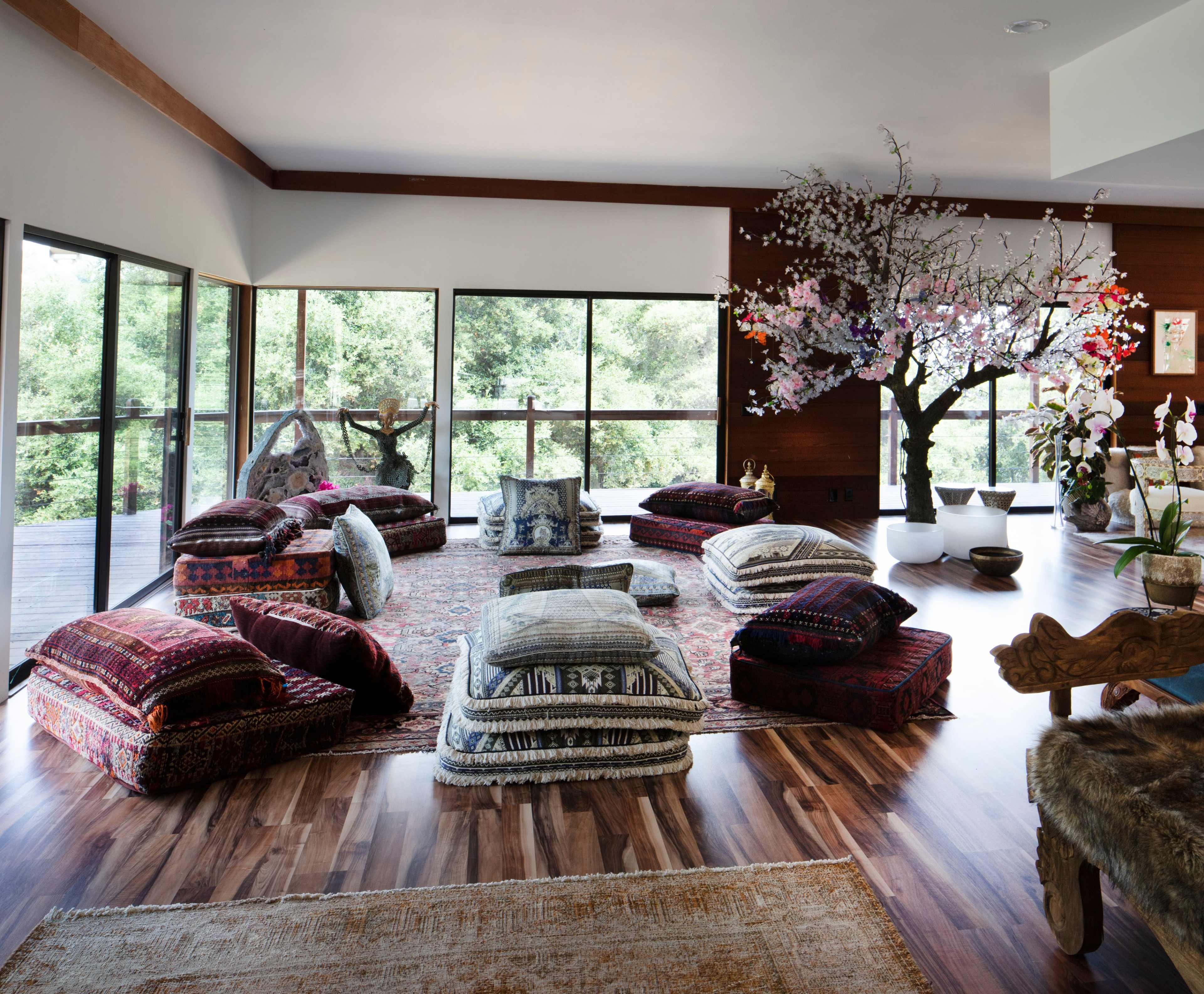 A spacious room is filled with colorful cushions arranged on a patterned rug, with large windows offering views of greenery and a decorative tree in one corner.
