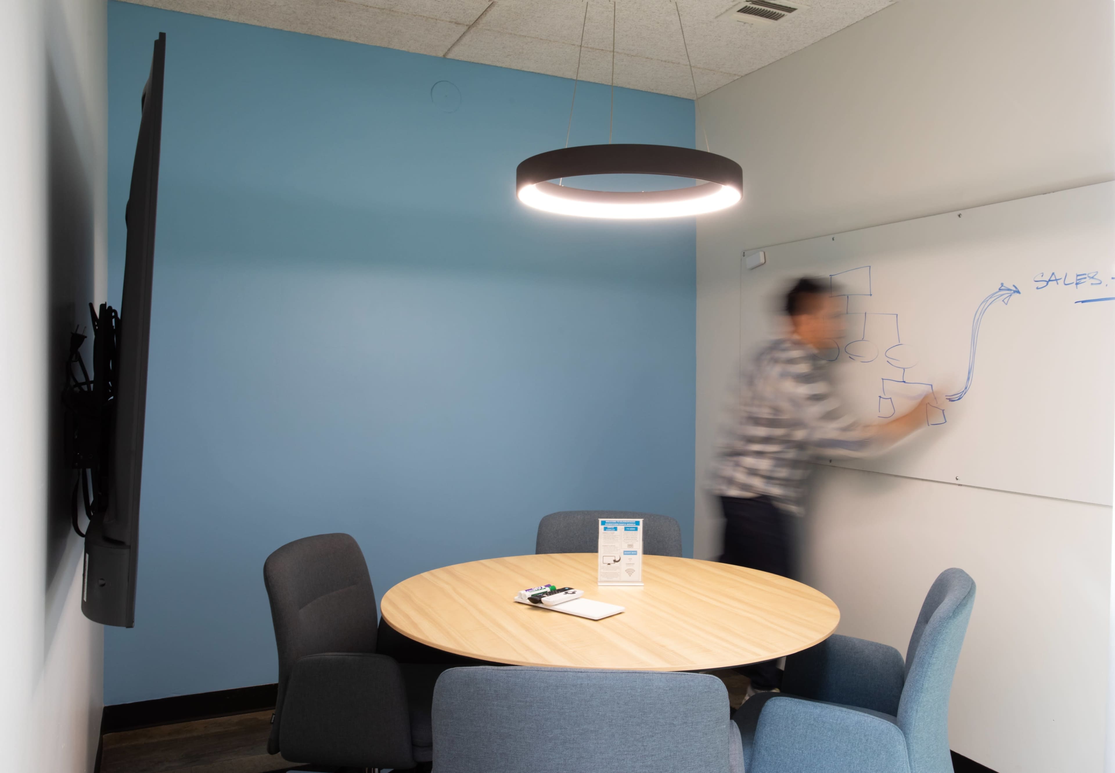A person is writing on a whiteboard in a small conference room with a circular table and a mounted TV.