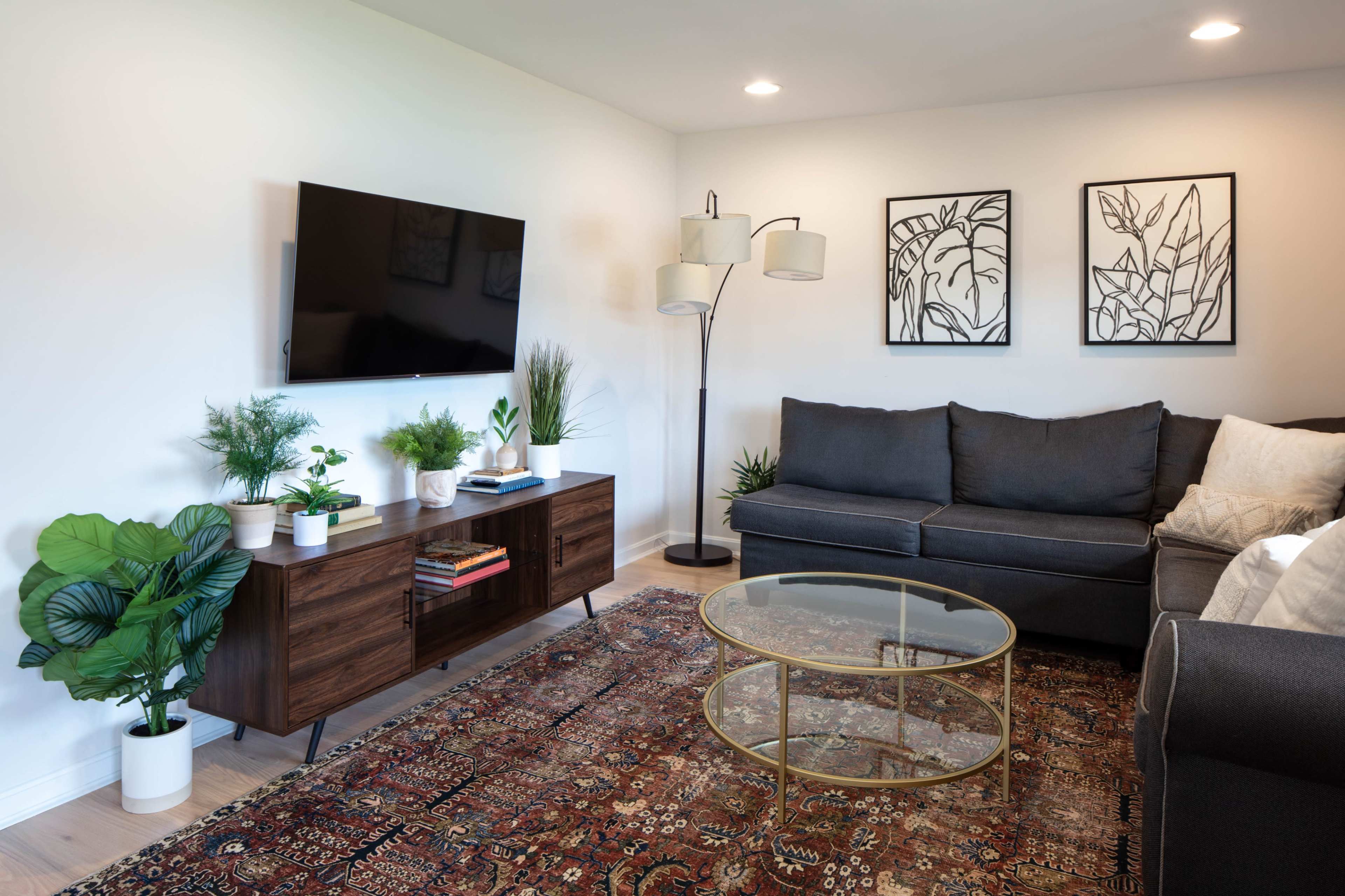 A modern living room features a gray sectional sofa, a wooden TV stand with plants, a round glass coffee table, and framed botanical artwork on the walls.