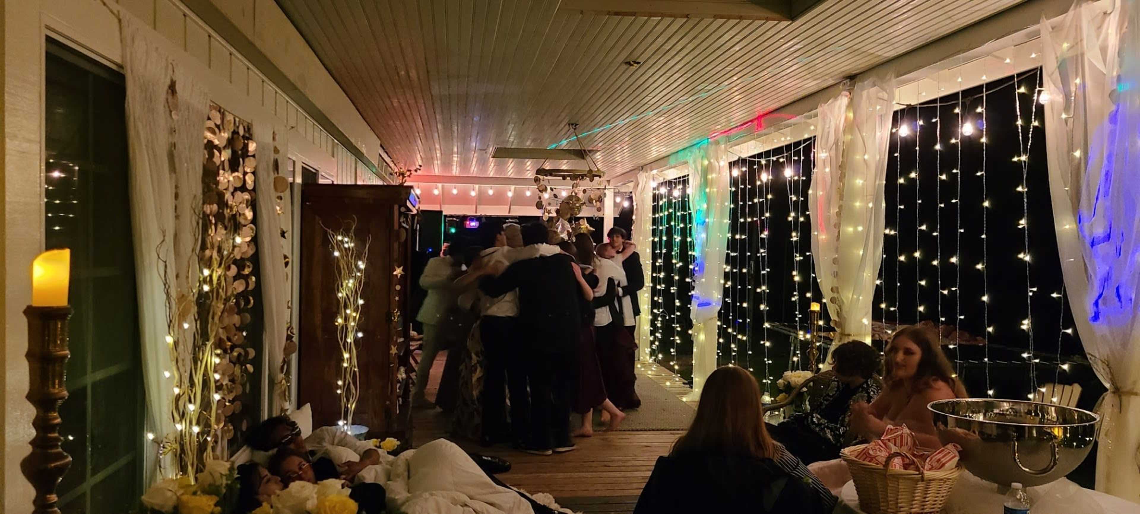A group of people gather and dance on a lit porch adorned with fairy lights and decorations.