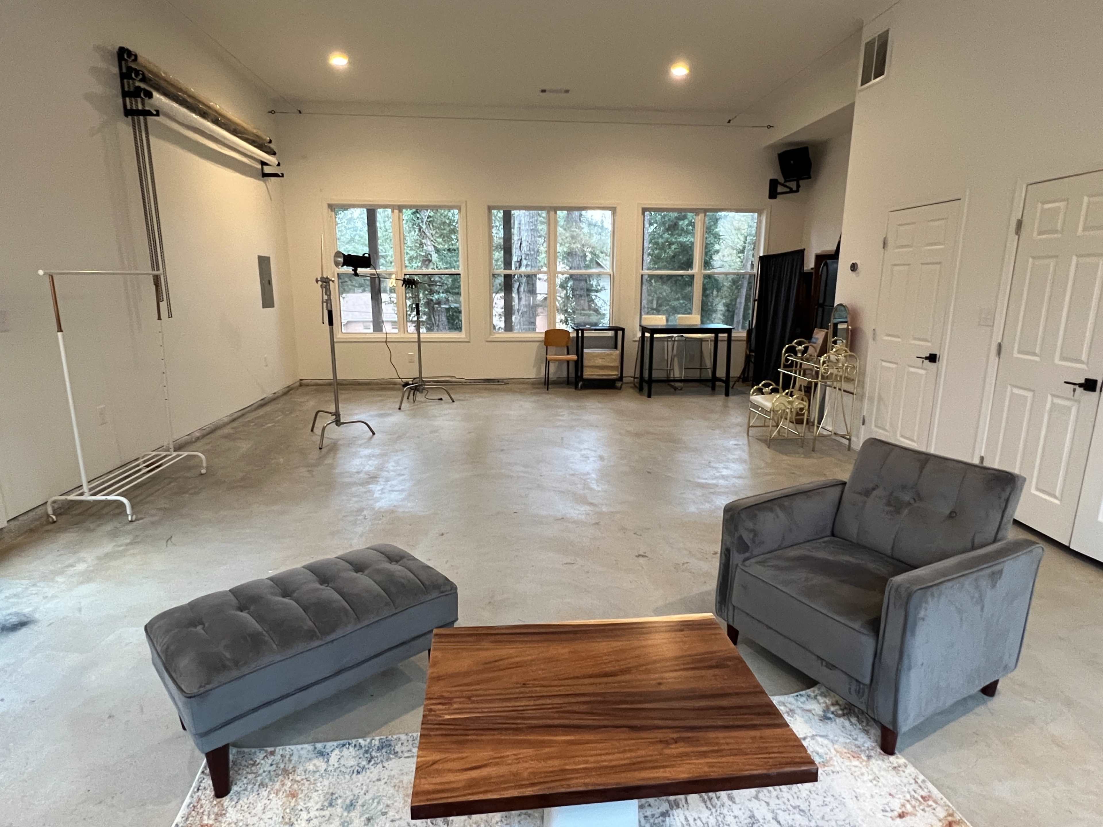 A spacious, empty room with concrete flooring, large windows, and minimal furniture, including two gray chairs and a wooden table.