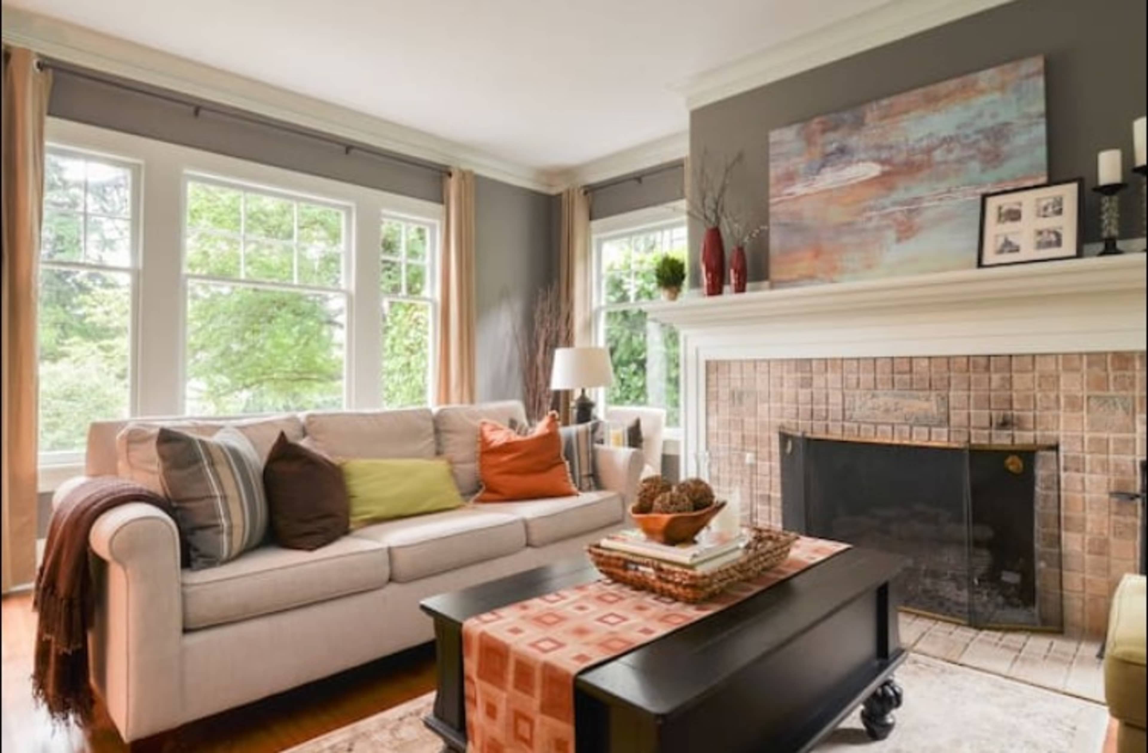 A cozy living room features a light-colored sofa with colorful pillows, a dark coffee table, and a tiled fireplace, surrounded by large windows letting in natural light.