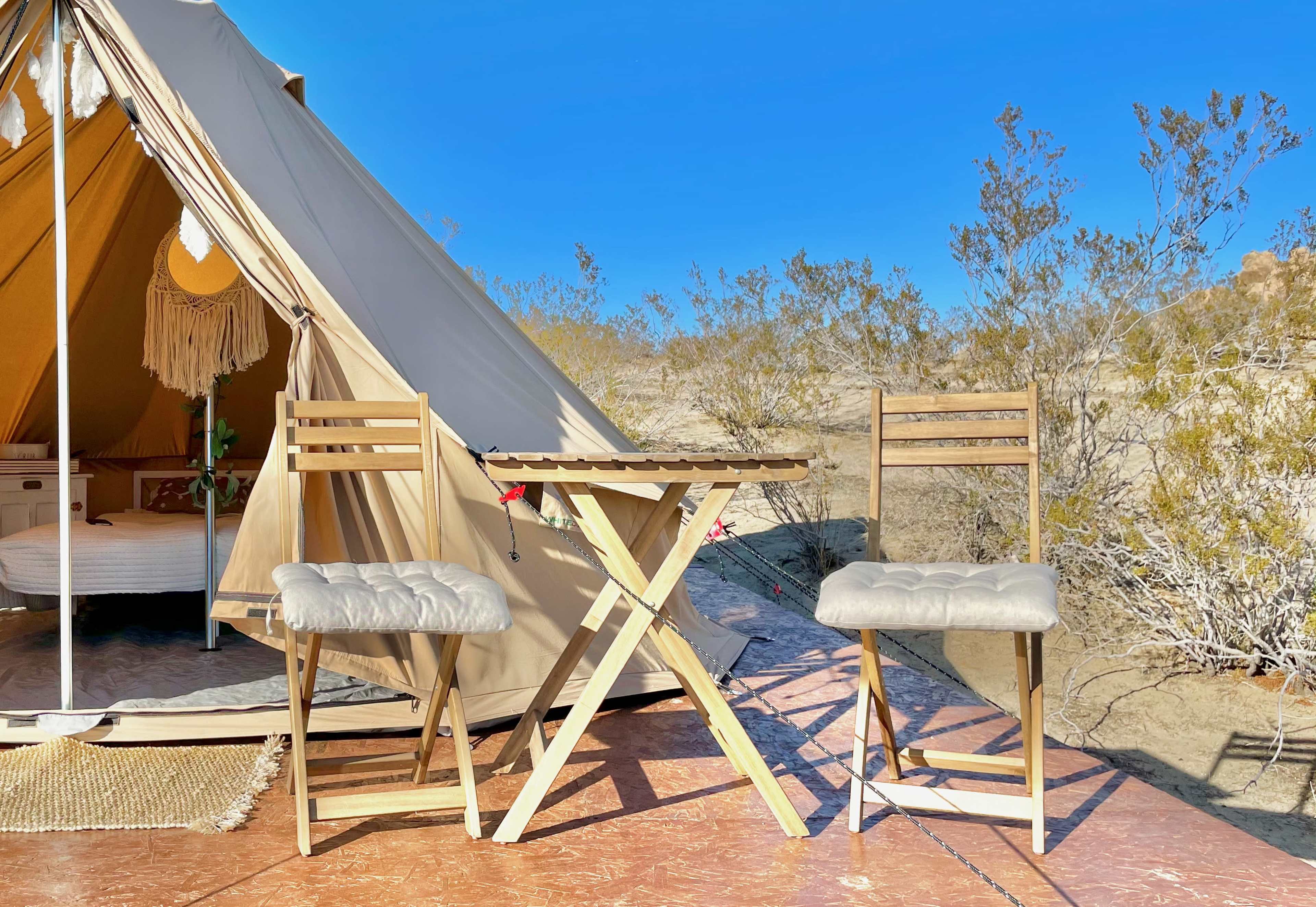 A wooden table and two chairs are set up in front of a canvas tent in a desert landscape.