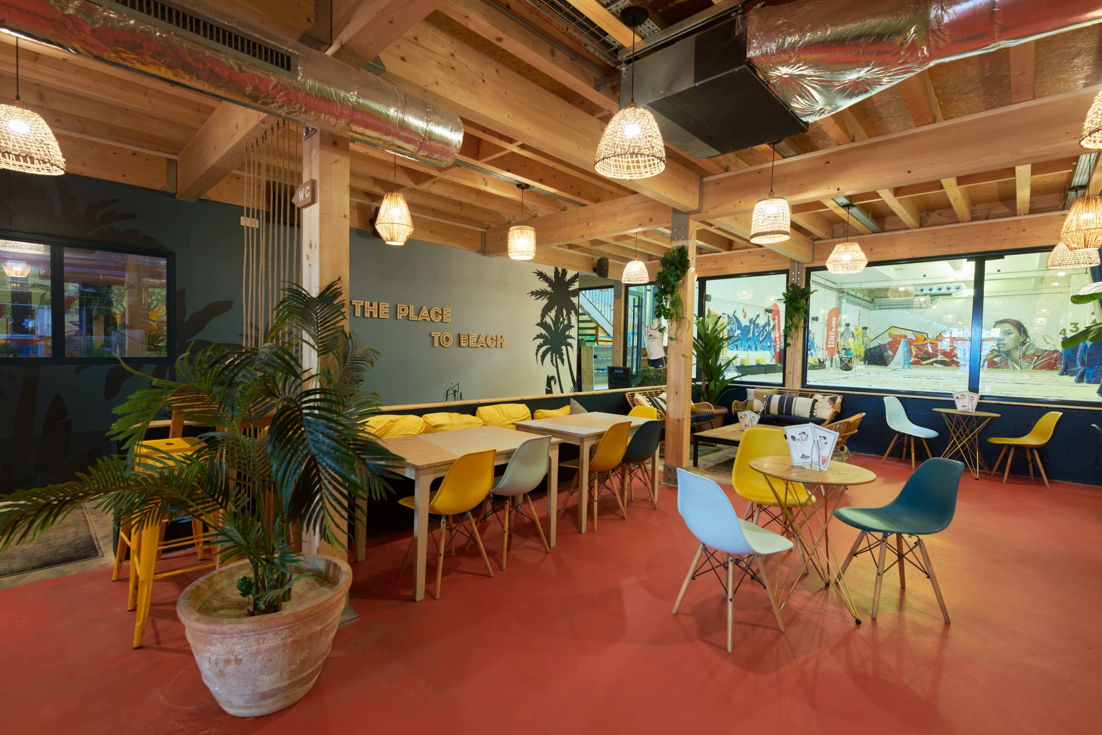 A modern indoor dining area with wooden beams, colorful chairs, and large windows featuring a view of a play area.