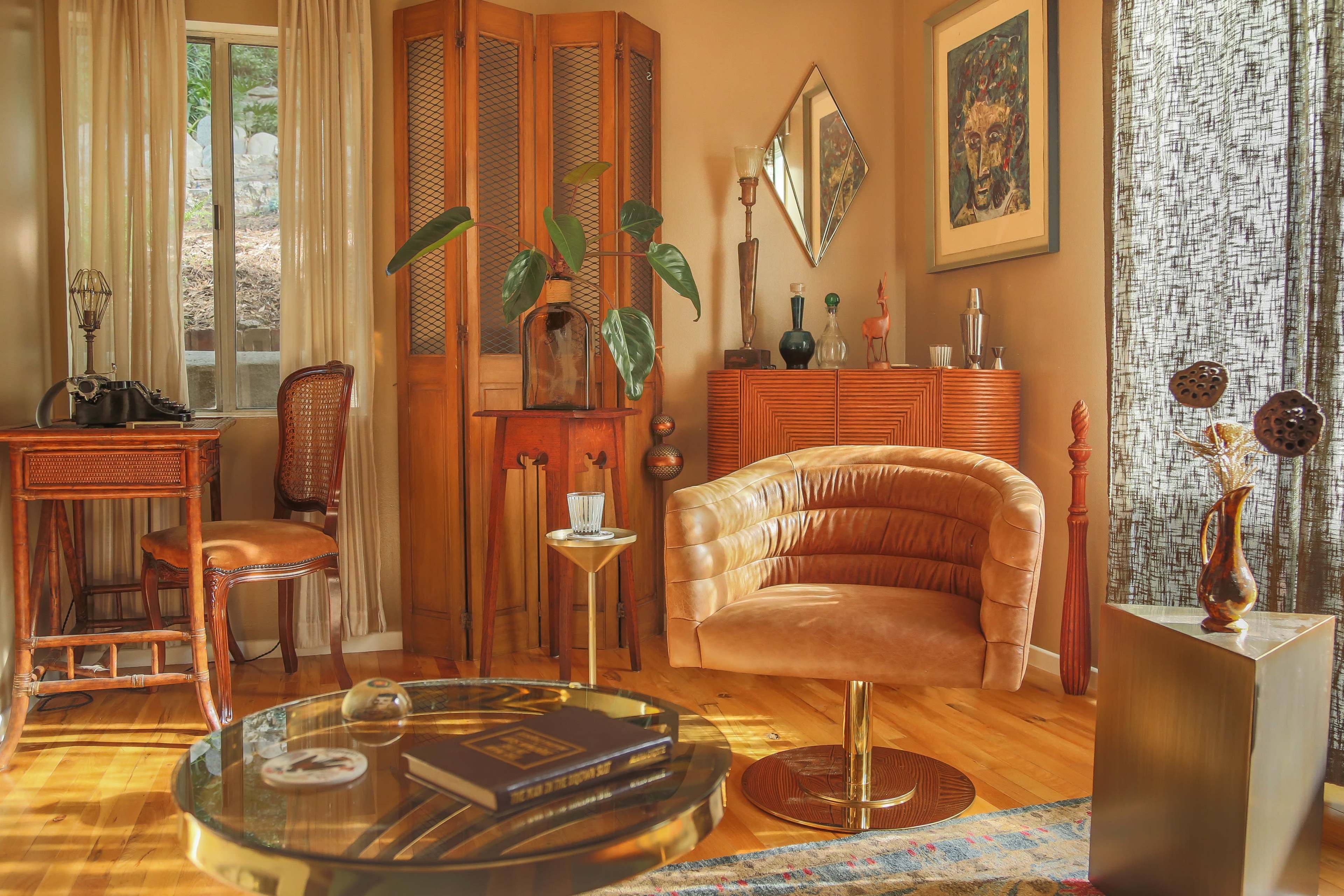The image depicts a stylish living room with mid-century furniture, including a plush armchair, a round coffee table, and a wooden cabinet, set against warm-toned walls and large windows.
