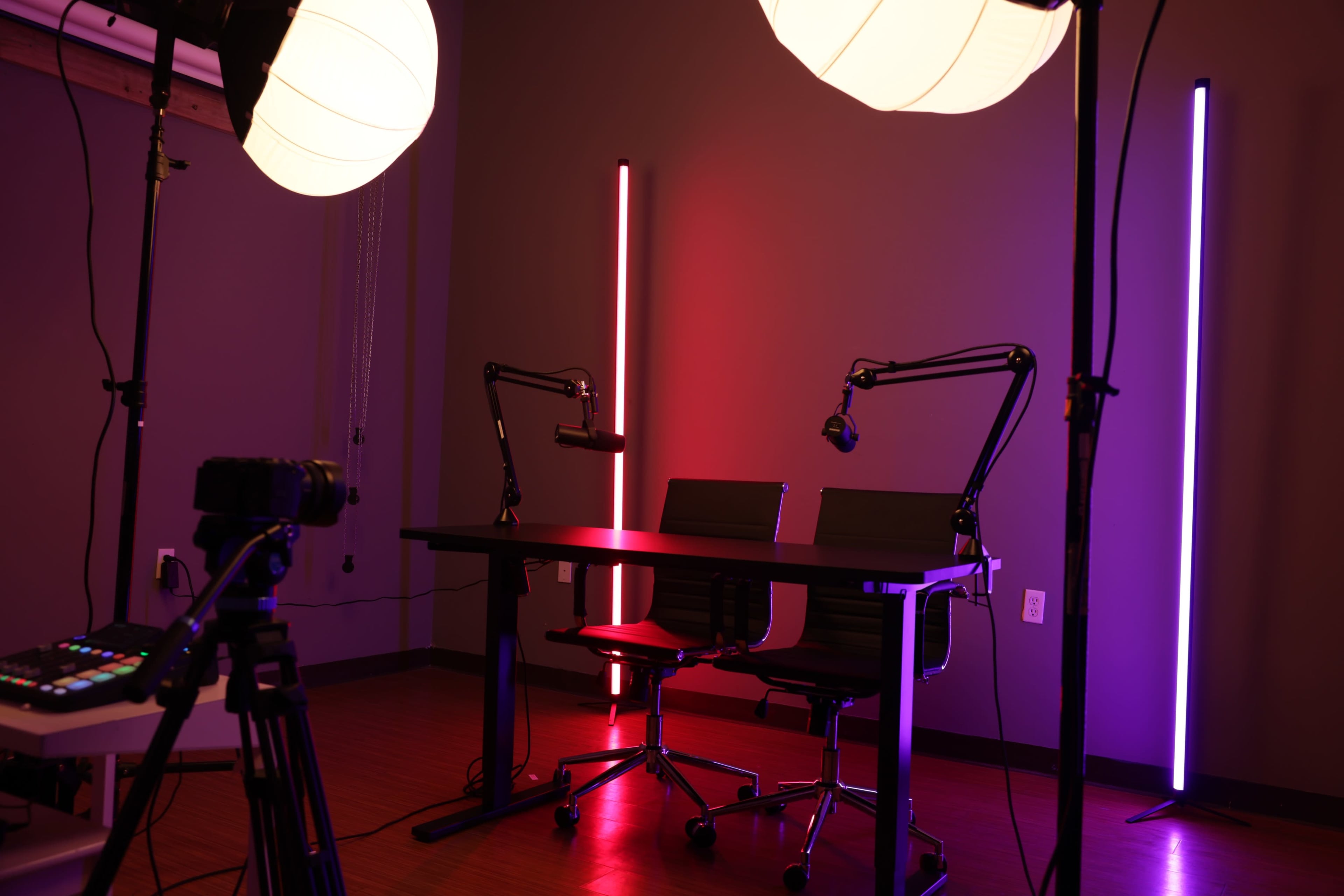 A modern studio setup features a table with two chairs, illuminated by colorful neon lights and equipped with microphones on adjustable arms.