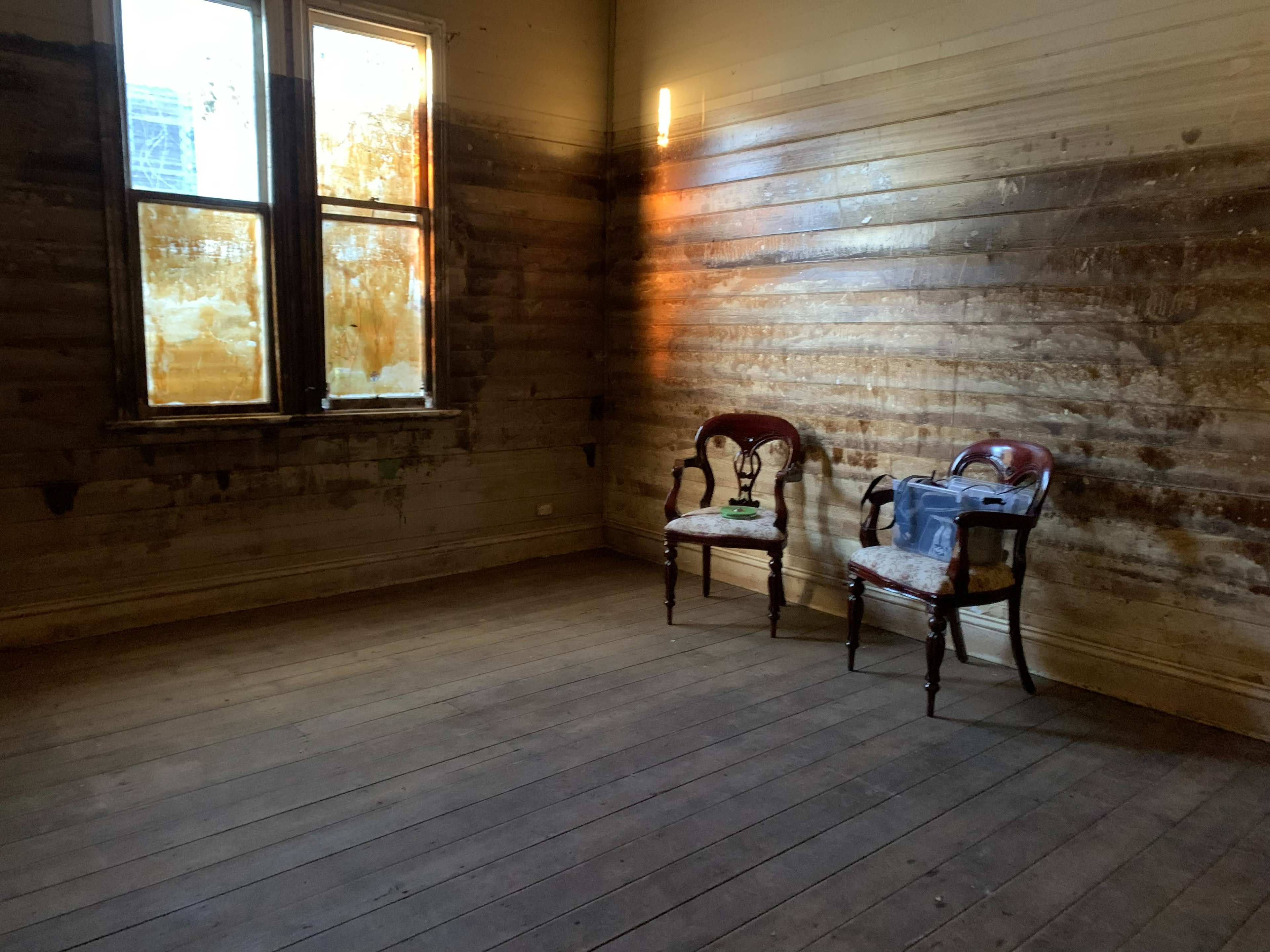 The image shows an empty room with two wooden chairs and a bare wooden floor, featuring peeling walls and two windows.