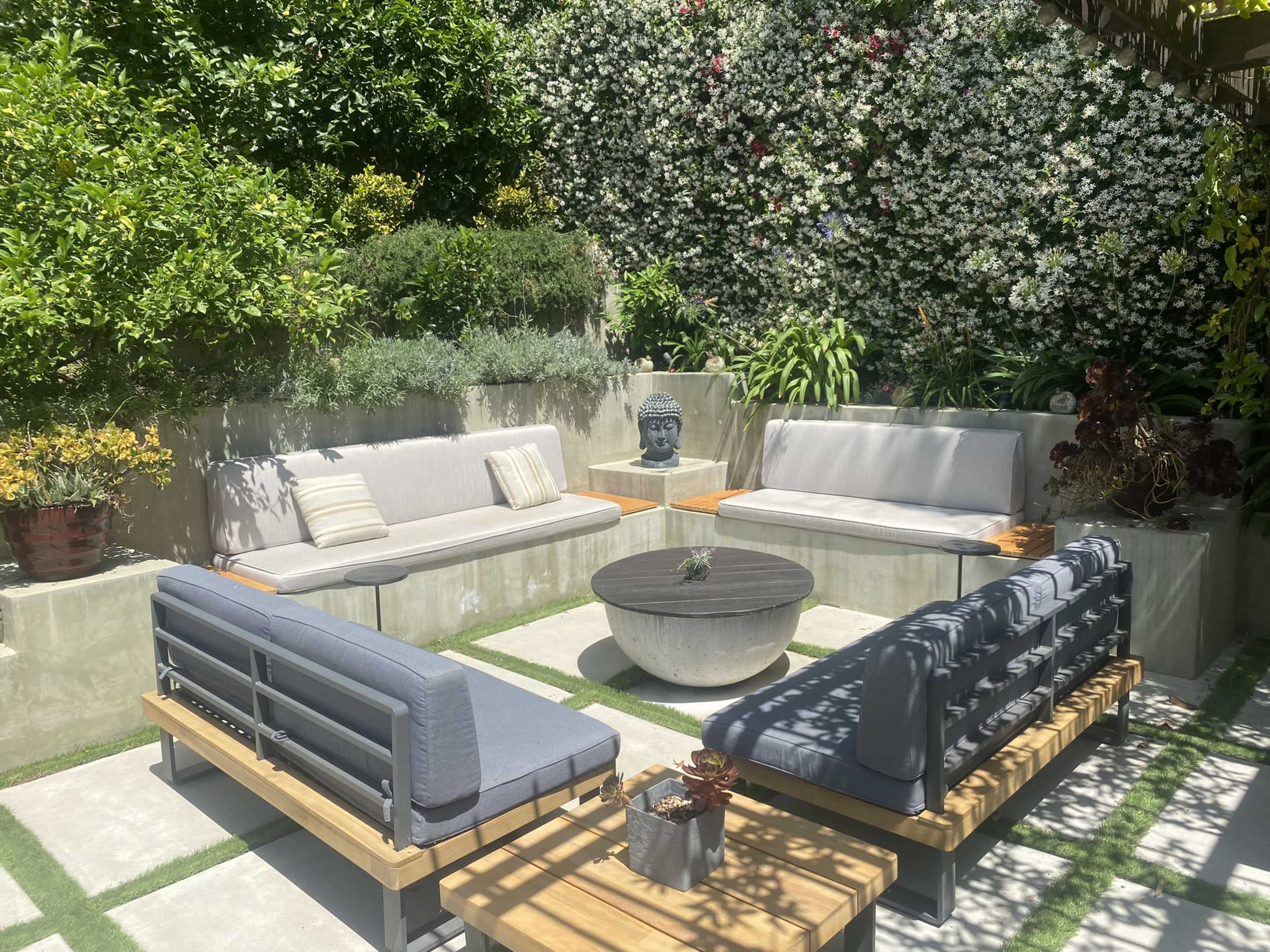 A modern outdoor seating area with gray sofas and a circular coffee table, surrounded by lush greenery and a wall covered in flowering plants.