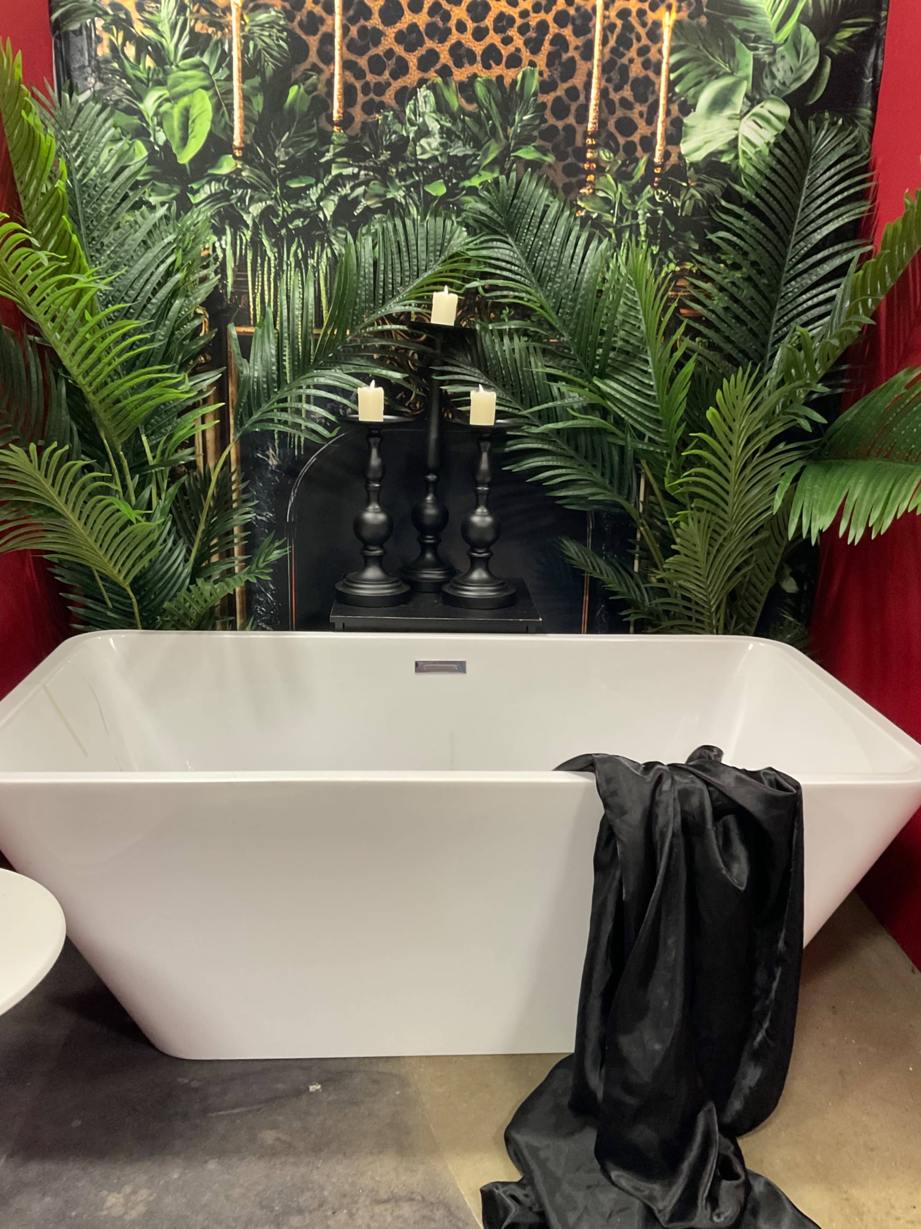 The image shows a modern white bathtub positioned against a vibrant green and black foliage backdrop, with three black candles on a stand amidst decorative plants.