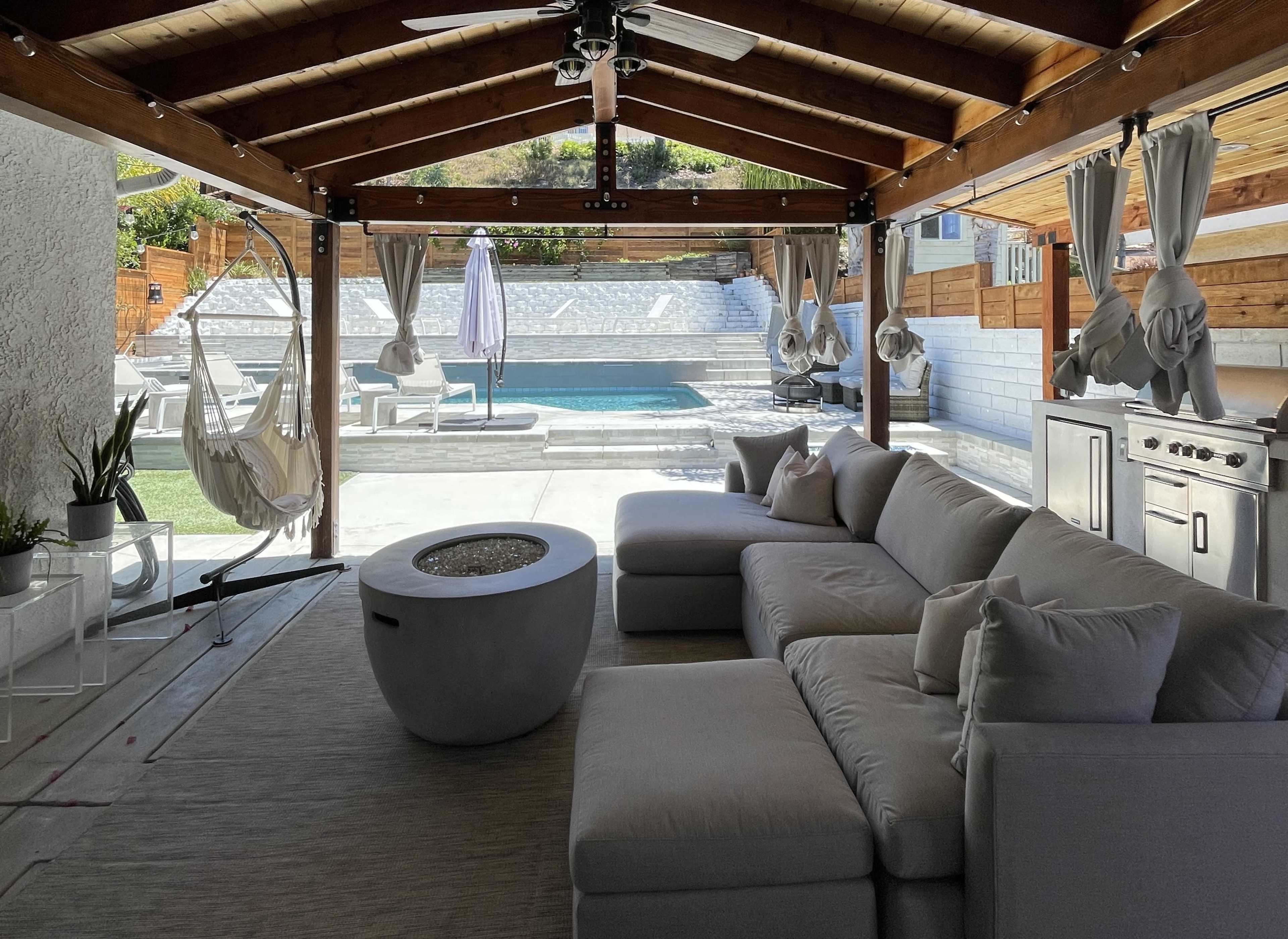 A covered patio features a sectional sofa, a modern fire pit, and a view of a swimming pool surrounded by lounge chairs.
