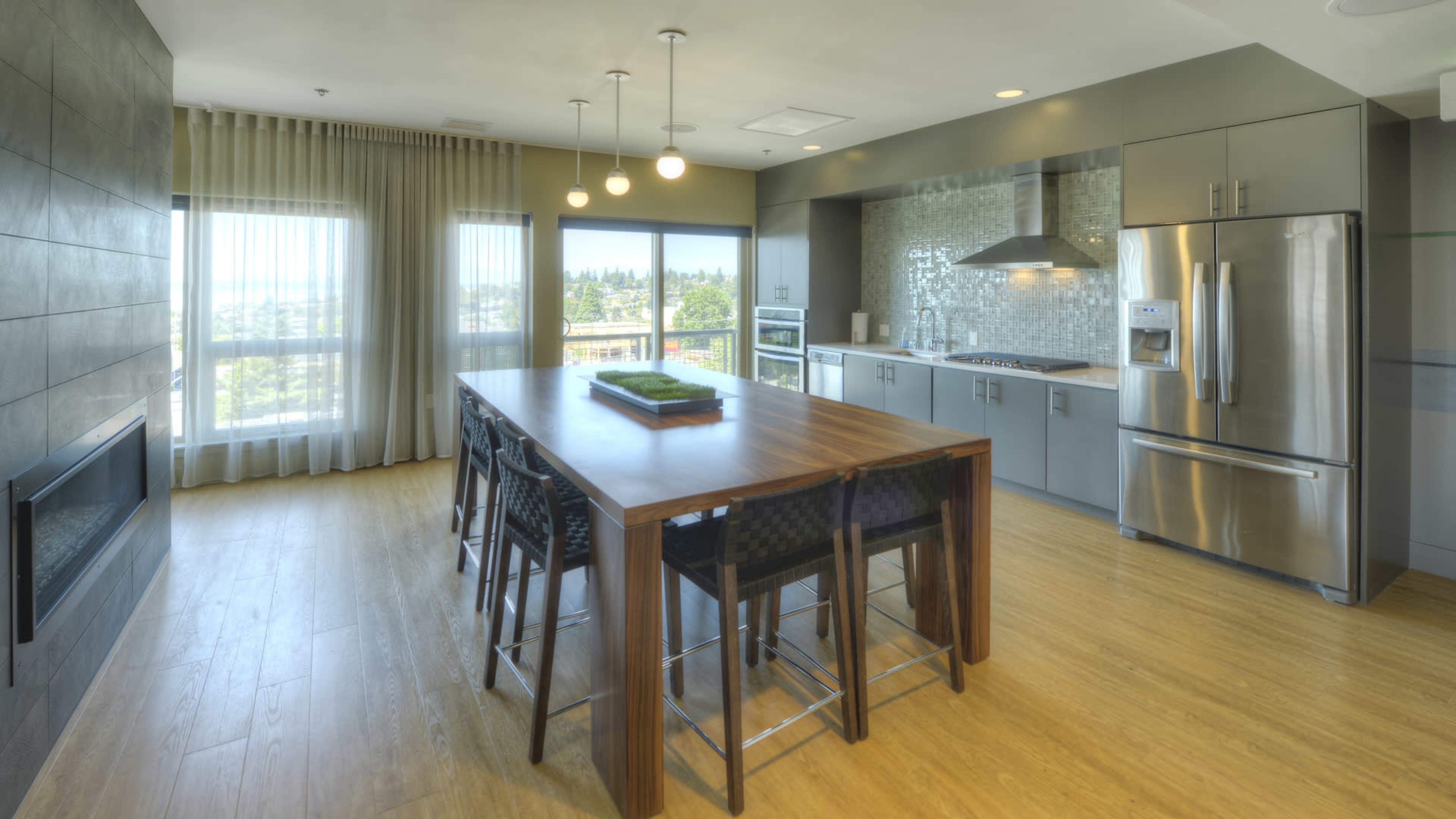 A modern kitchen and dining area features a large wooden table with high-backed chairs, stainless steel appliances, and floor-to-ceiling windows with sheer curtains.