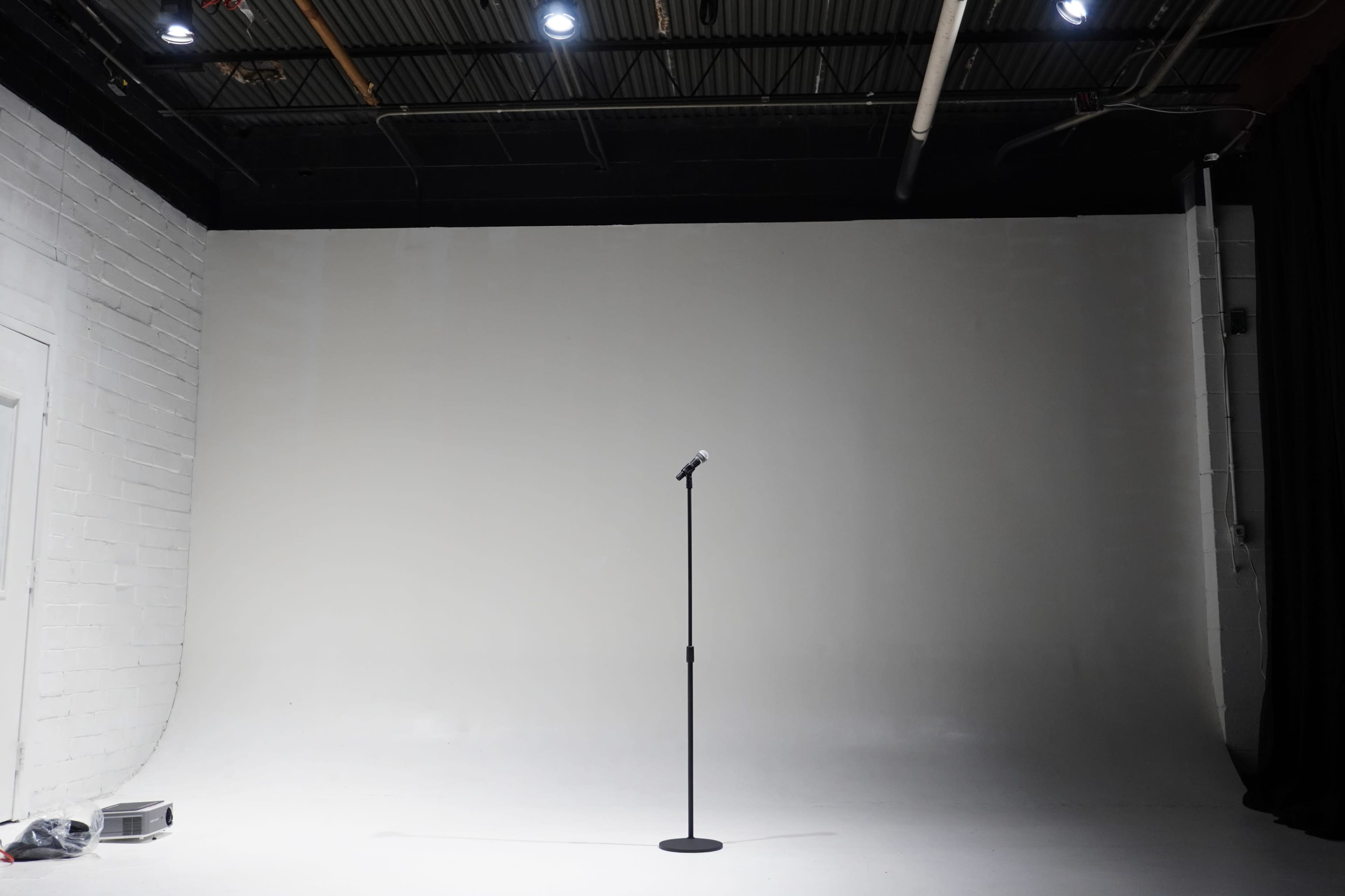 The image shows a minimalist stage with a single microphone stand centered in front of a plain white backdrop.