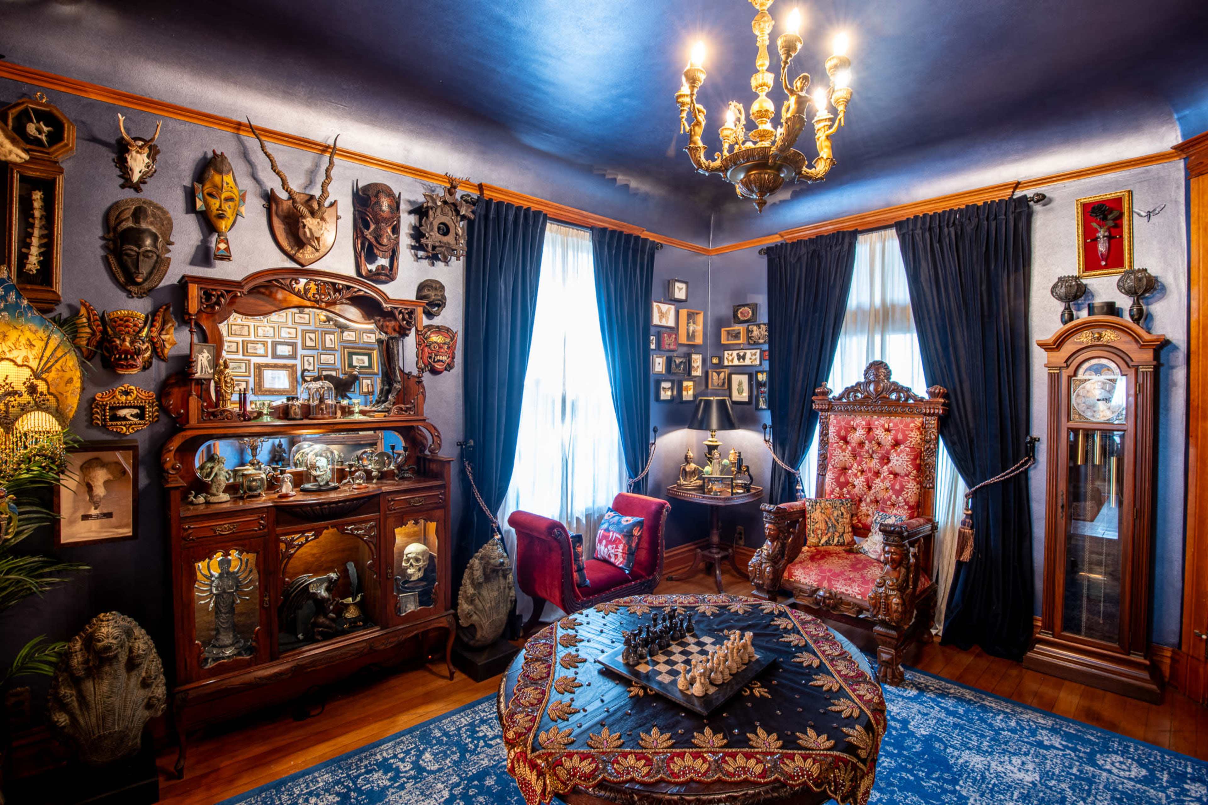 The room features ornate furniture and decor, including a large chandelier, a chess set on a central table, and walls adorned with various masks and framed photos.