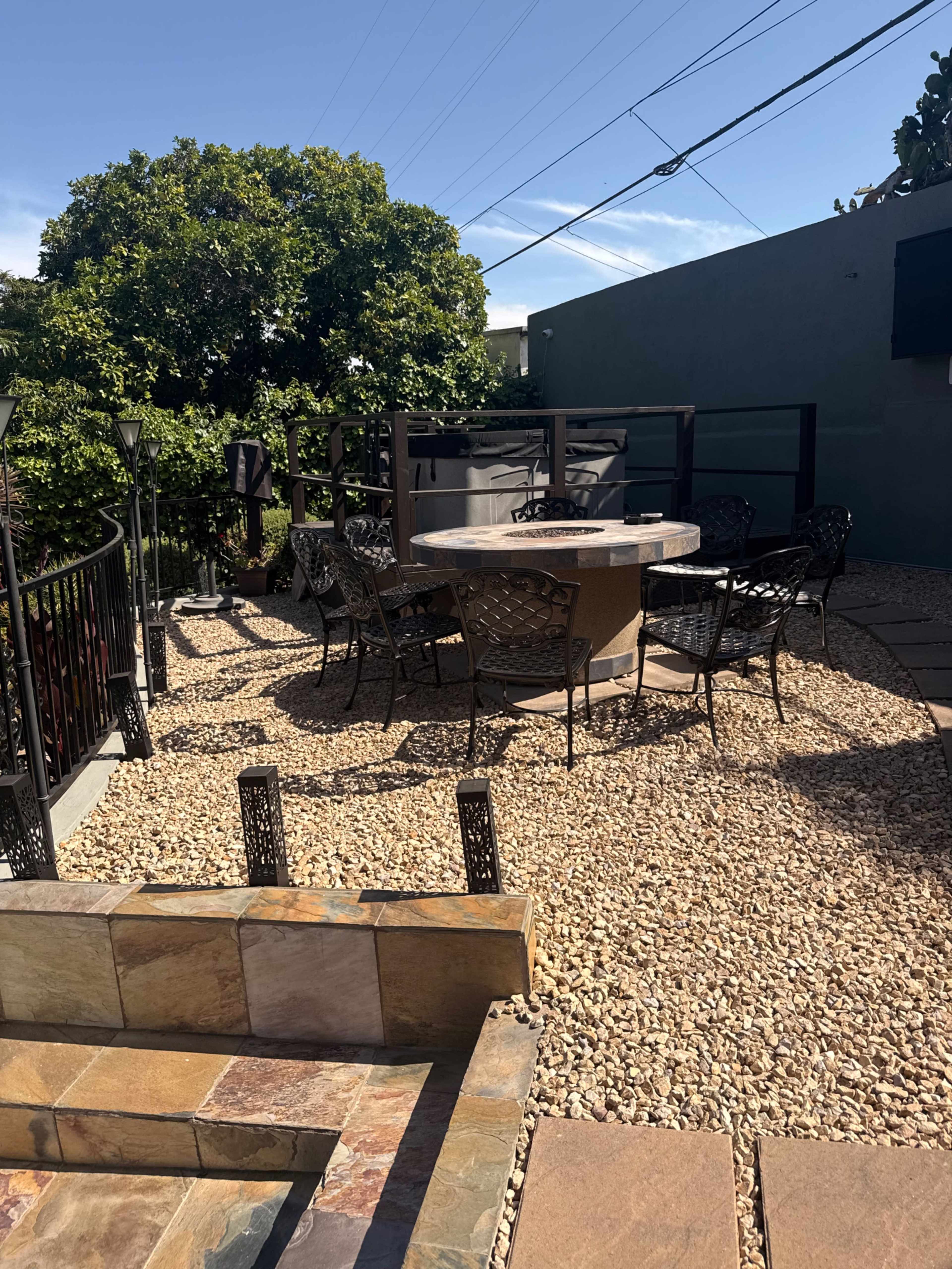 The image shows an outdoor patio area with a round stone table surrounded by metal chairs, situated on a gravel surface with a railing and trees in the background.
