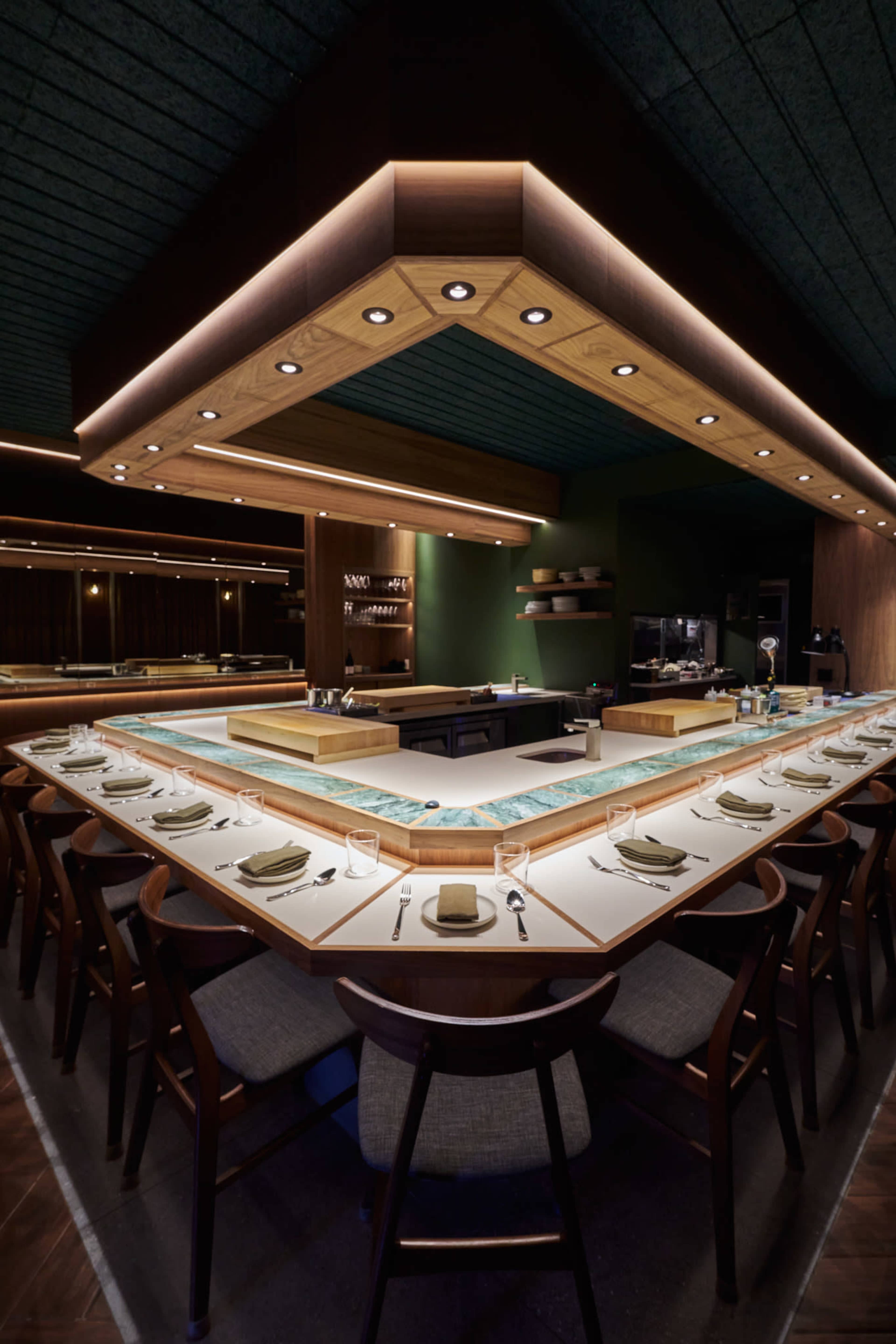 The image showcases a modern restaurant interior featuring a large sushi bar with an illuminated wooden canopy and a row of dining chairs arranged around a central countertop.