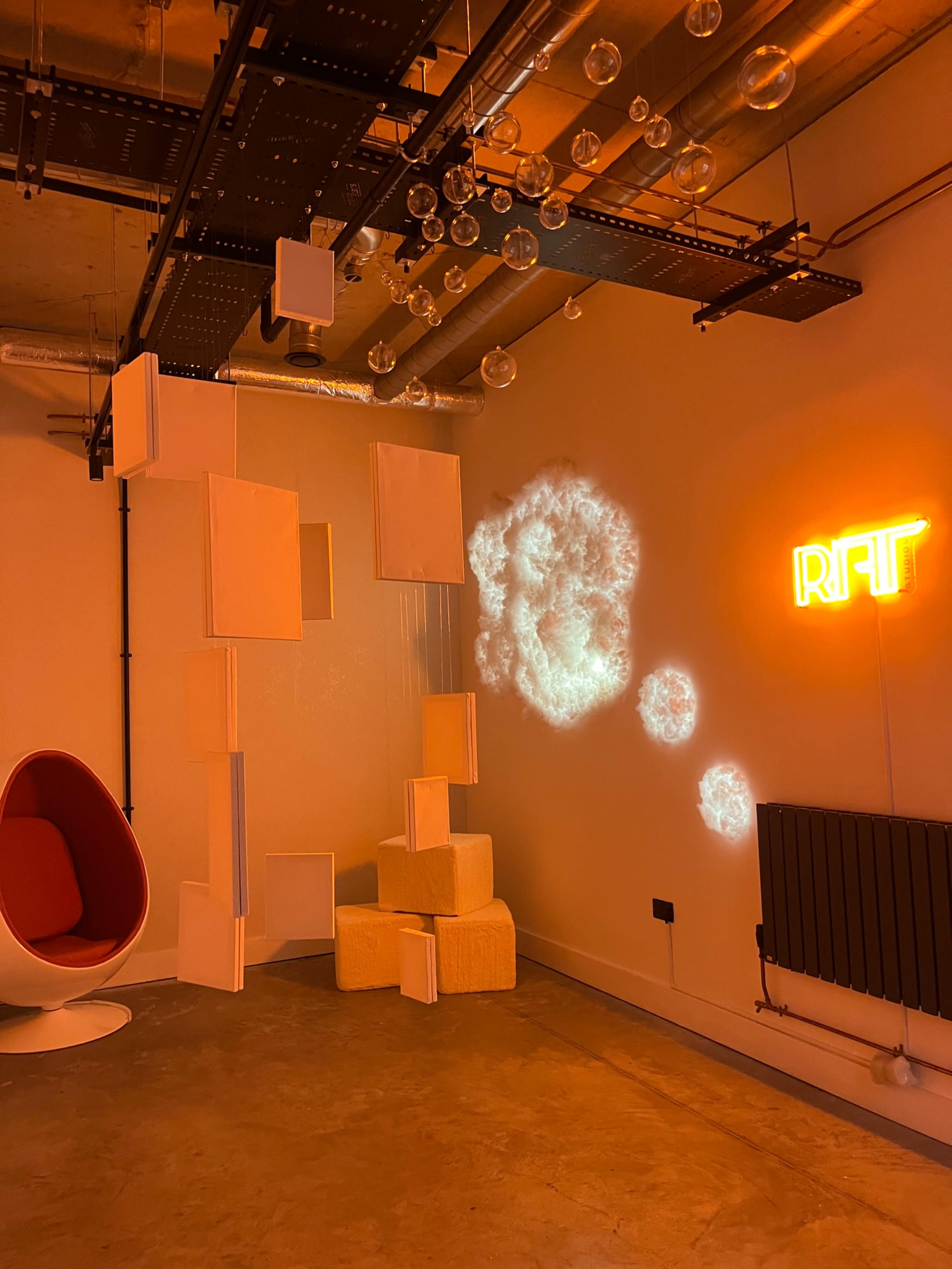 The image shows a modern indoor space featuring a red egg-shaped chair, abstract white sculptures, a neon sign that reads "RFT," and decorative hanging glass orbs against a light-colored wall.