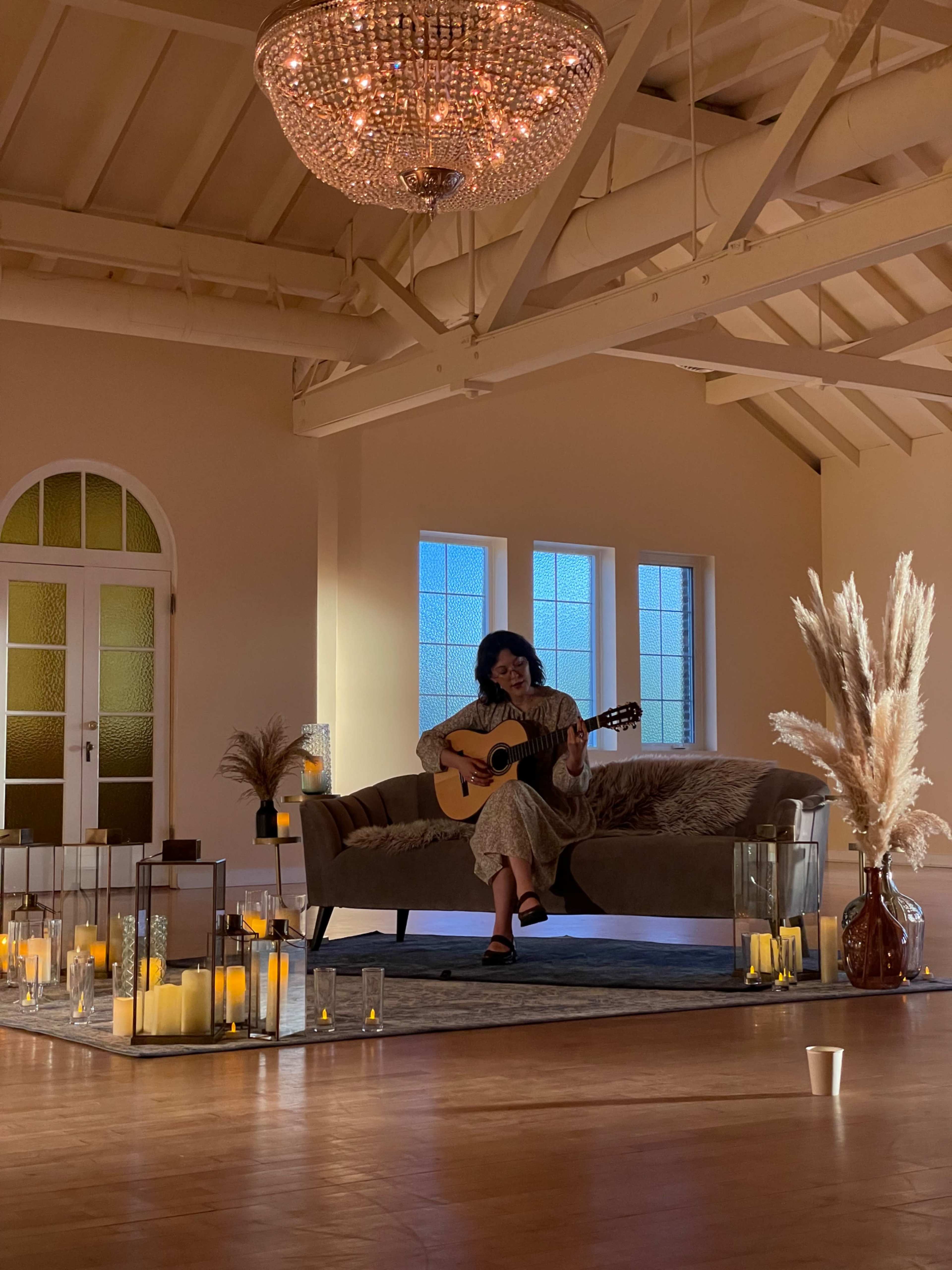 A person sits on a sofa in a softly lit room, playing an acoustic guitar surrounded by candles and decorative plants.