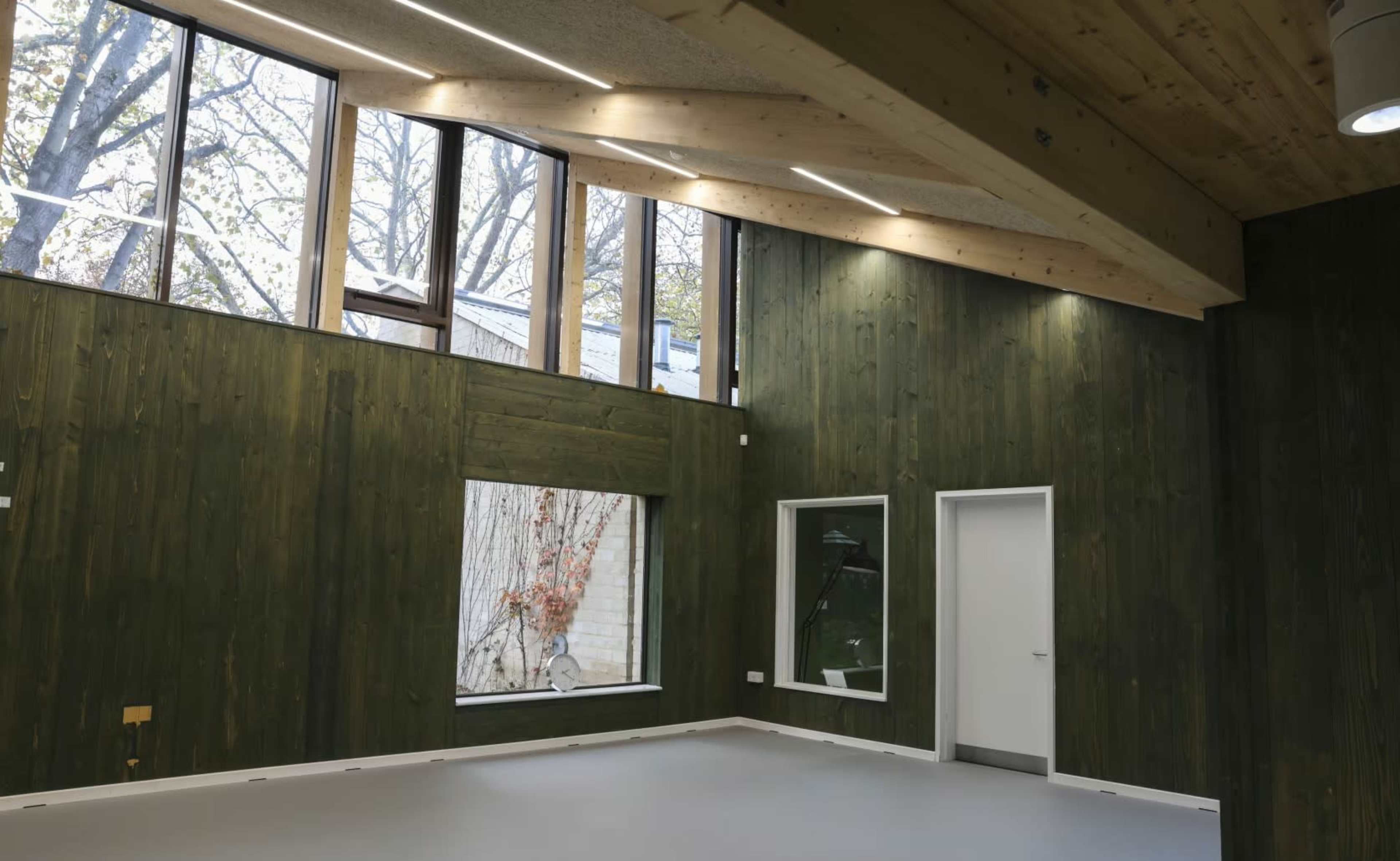 The image shows a modern interior space with green wooden walls, large windows allowing natural light, and minimal furnishings.