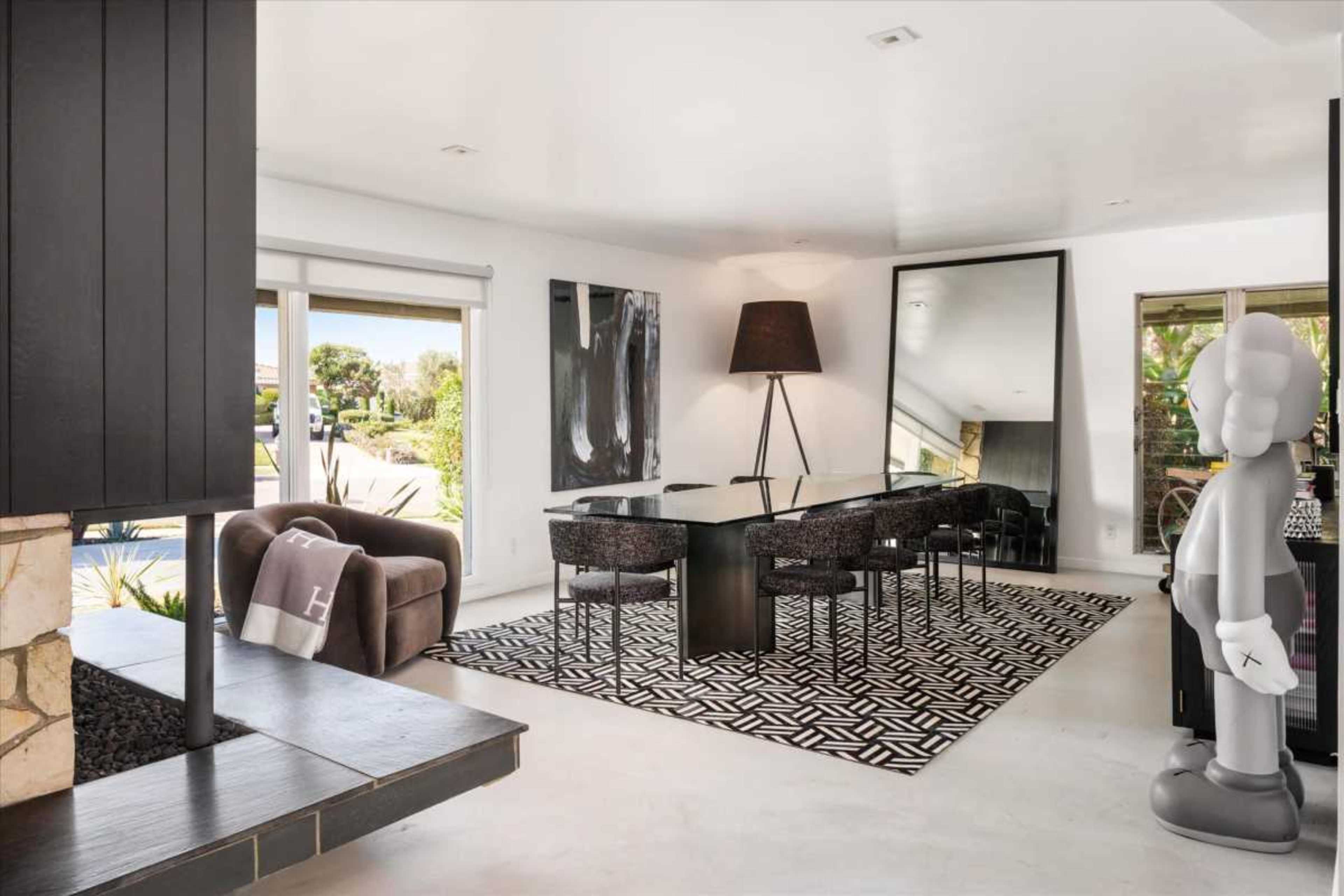 The scene shows a modern dining area with a black glass table surrounded by dark chairs, a large floor lamp, and a decorative sculpture, all positioned against a backdrop of large windows and contemporary art pieces.