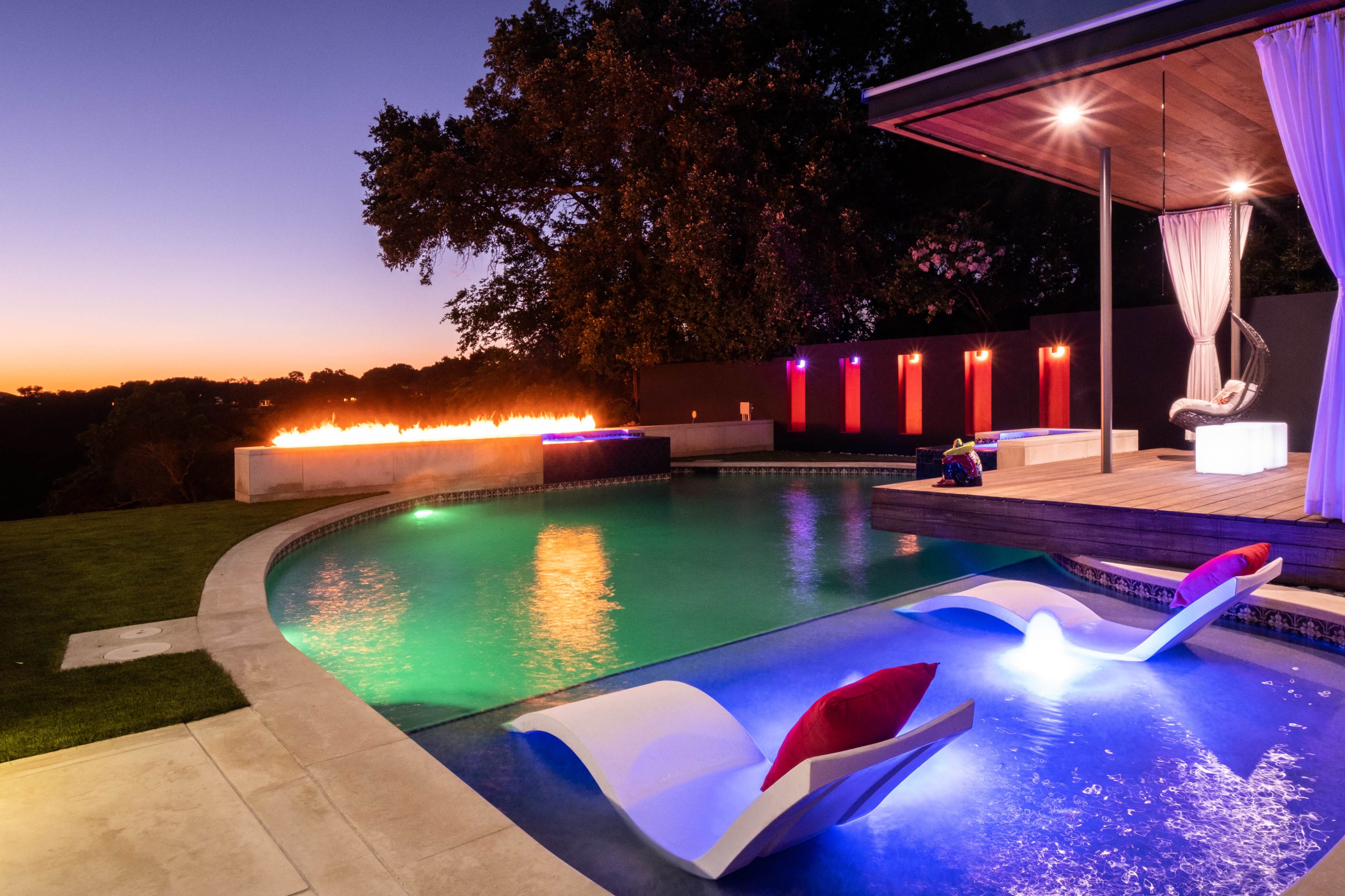 A modern swimming pool area features illuminated seating and a fire display against a twilight sky.