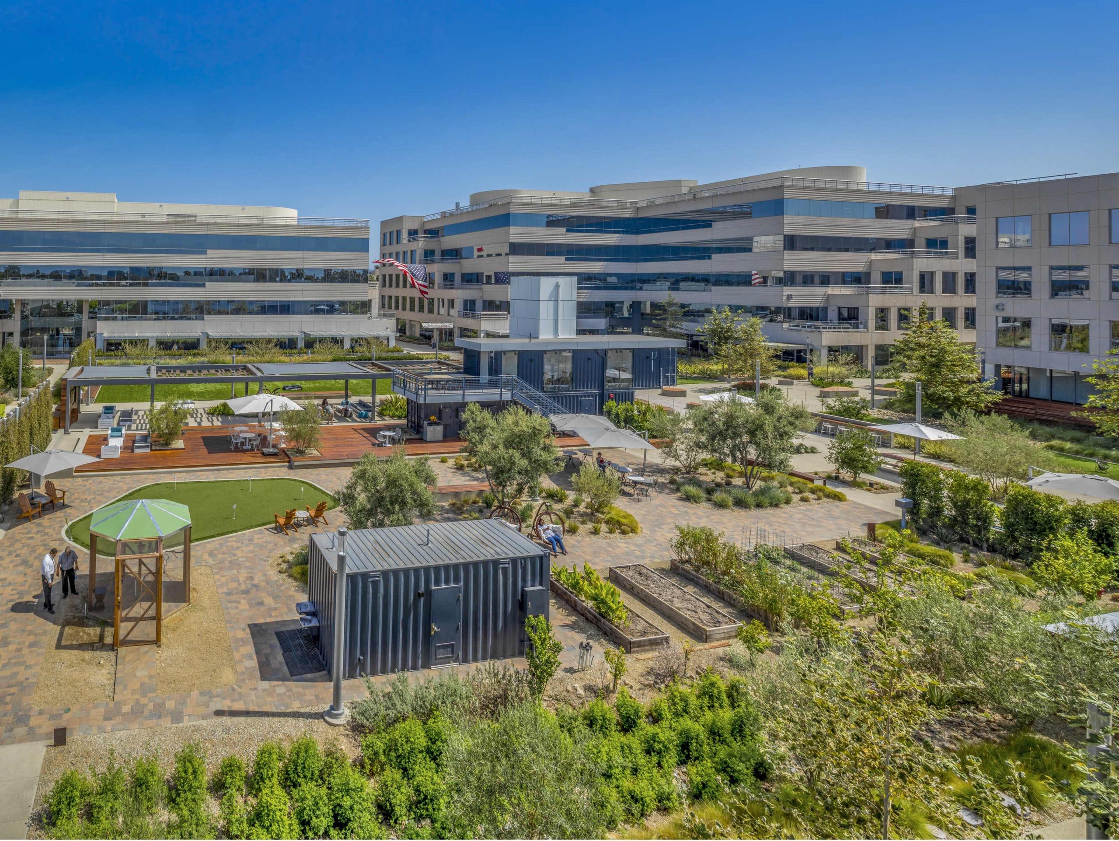 The image shows a large office complex with landscaped outdoor seating areas, walkways, and a playground in the foreground.