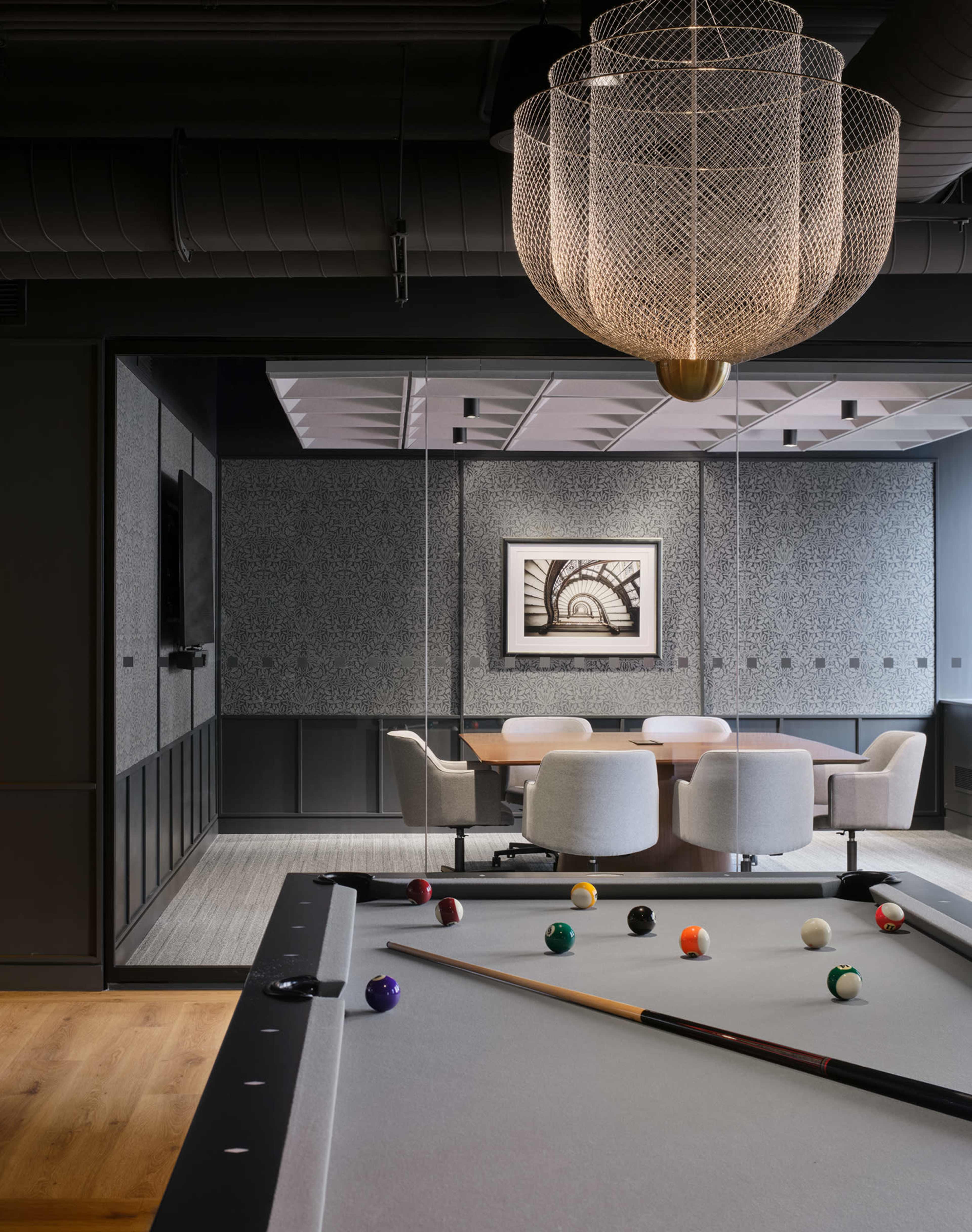 A billiards table is positioned in the foreground, while a glass-walled meeting room with a round table and upholstered chairs is visible in the background.