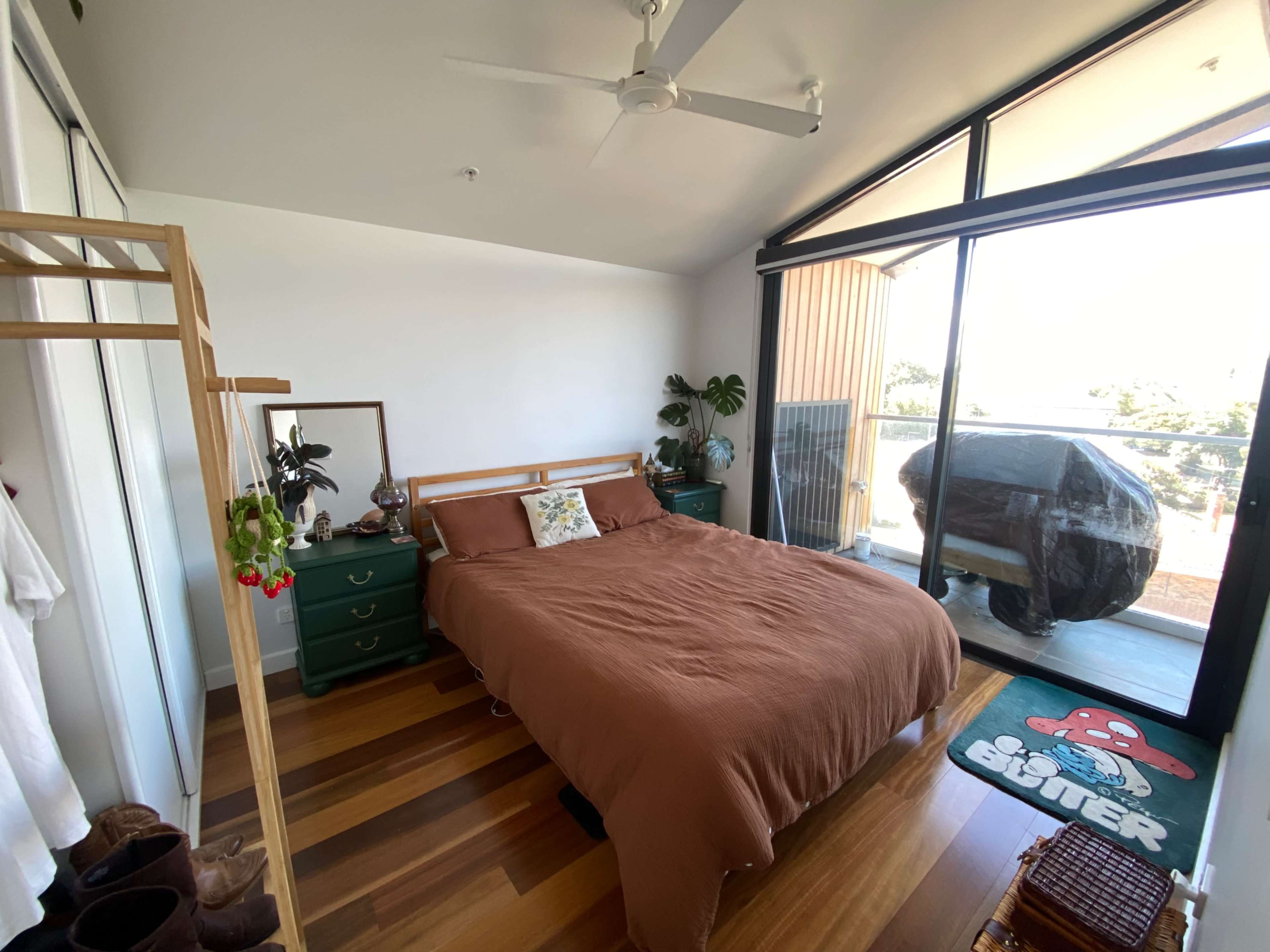 The bedroom features a brown bedspread, wooden flooring, a plant beside the window, and a covered outdoor grill visible on the balcony.
