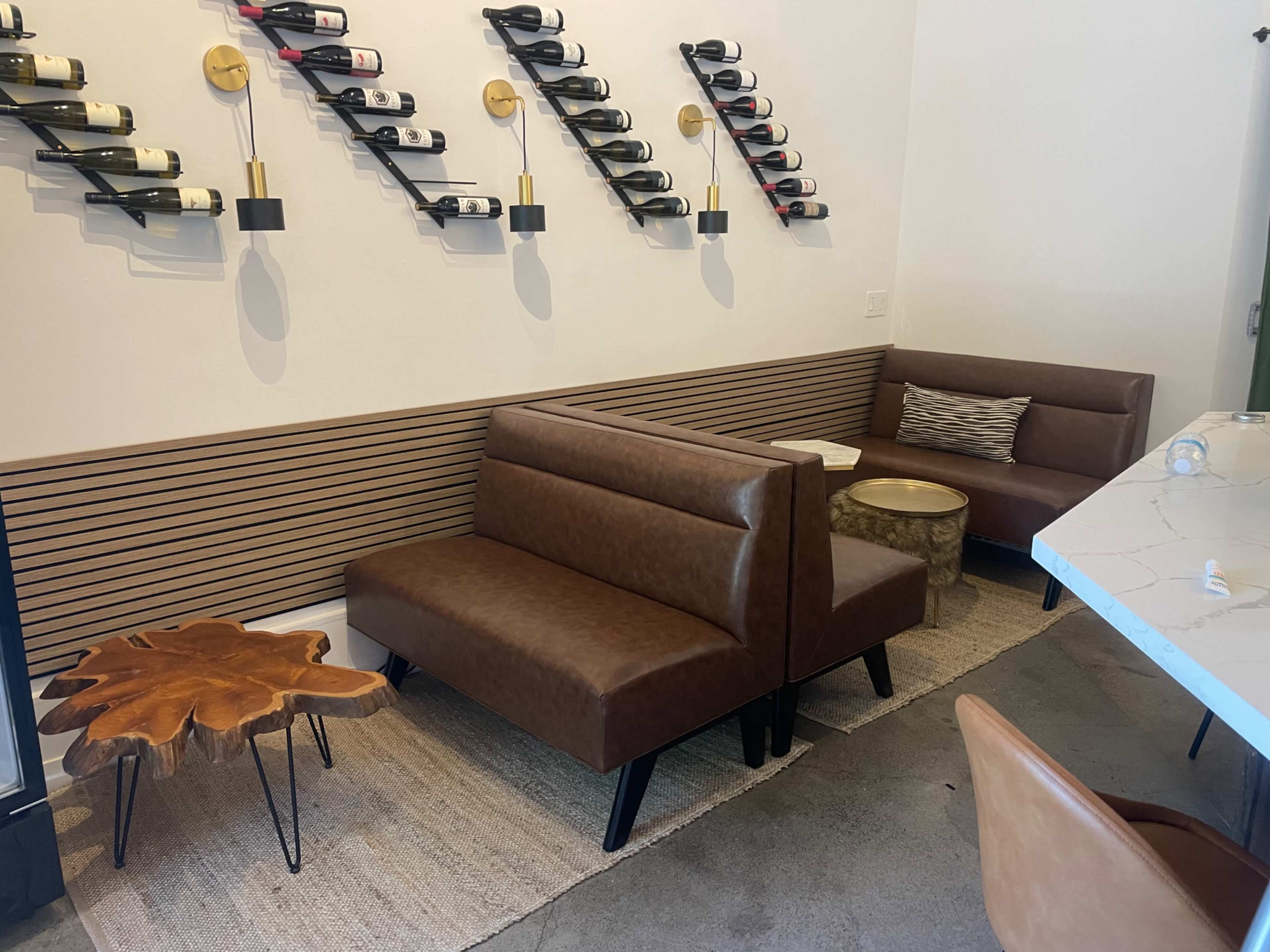 The image shows a modern seating area with brown leather benches, a small wooden table shaped like a leaf, and wall-mounted wine racks in a neutral-toned space.