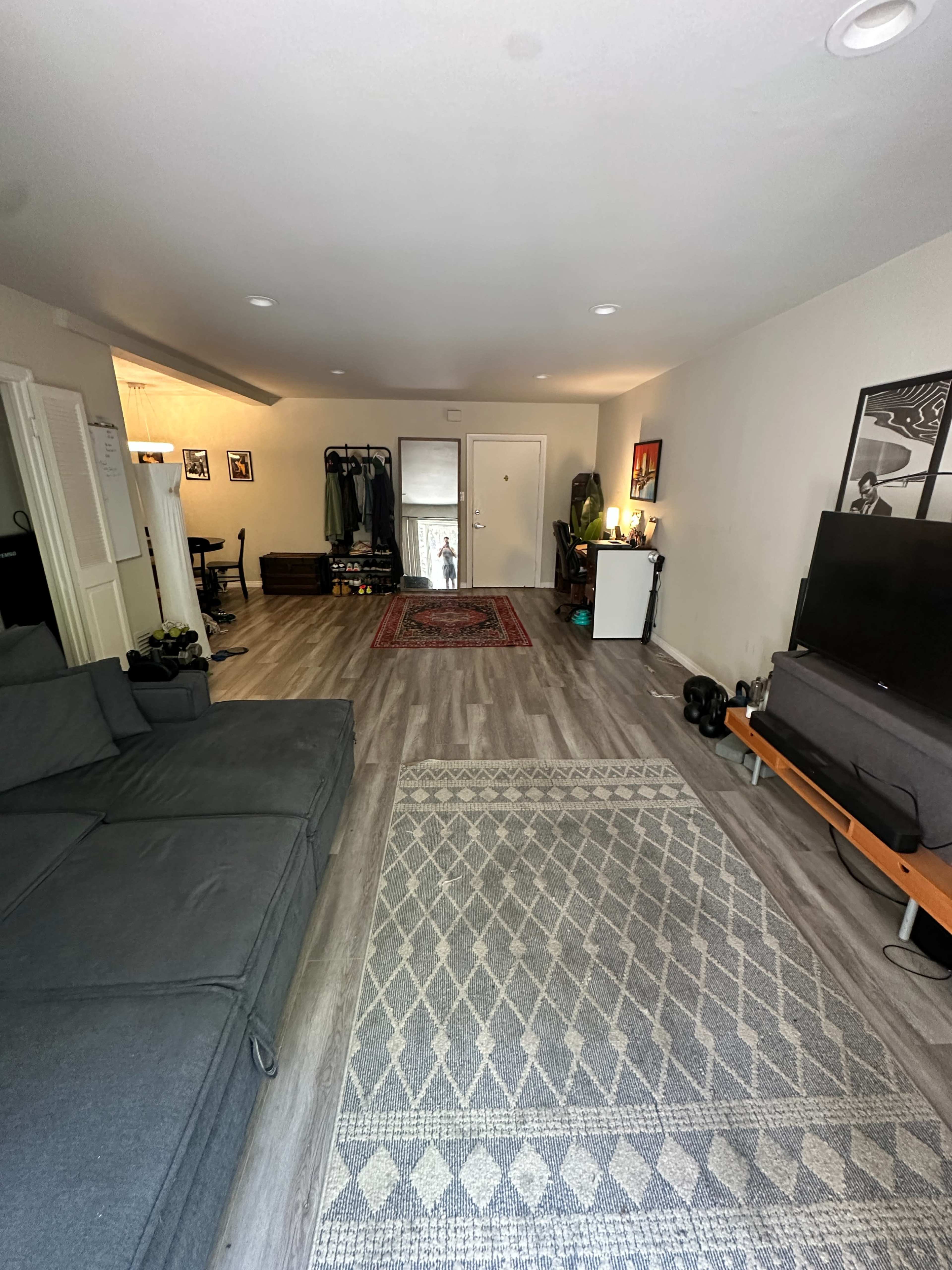 The image shows a spacious living room with a gray sectional sofa, a TV on a stand, and various furnishings along the walls, leading to a doorway and another room.