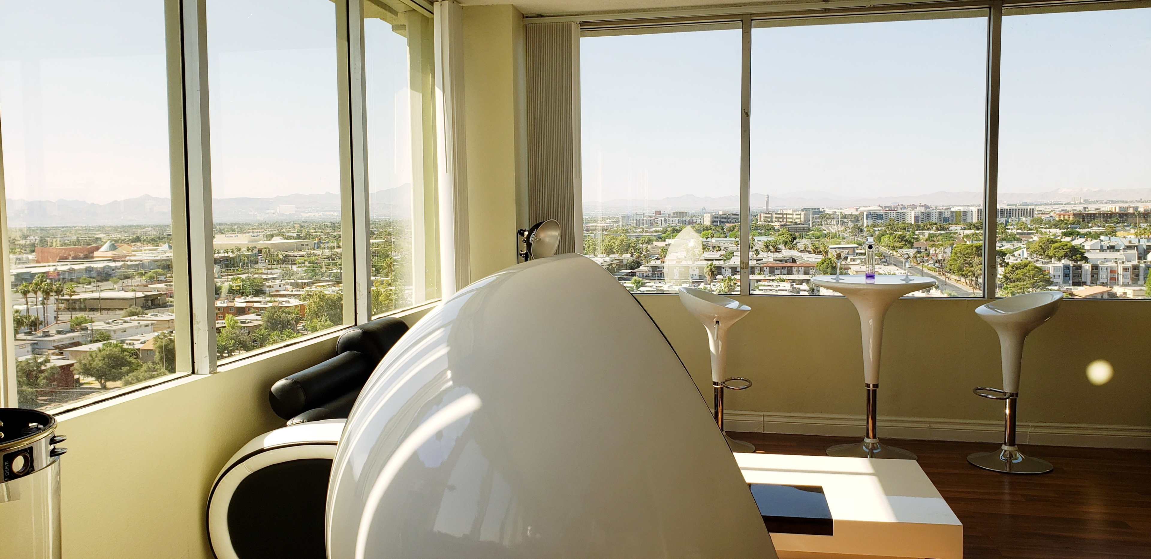 A modern room with large windows offering a view of a cityscape, featuring a sleek white seating area and bar-style stools.
