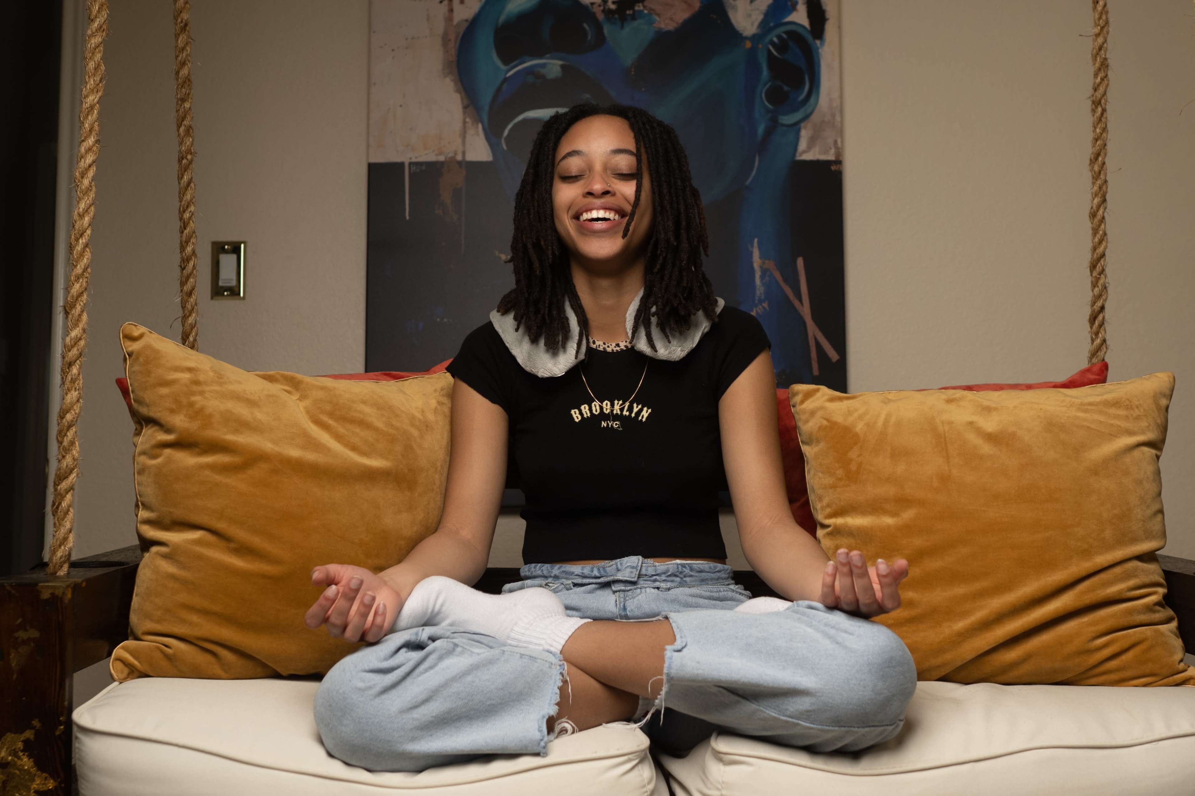 A young woman is sitting cross-legged on a couch, smiling while practicing meditation in front of a colorful wall art.