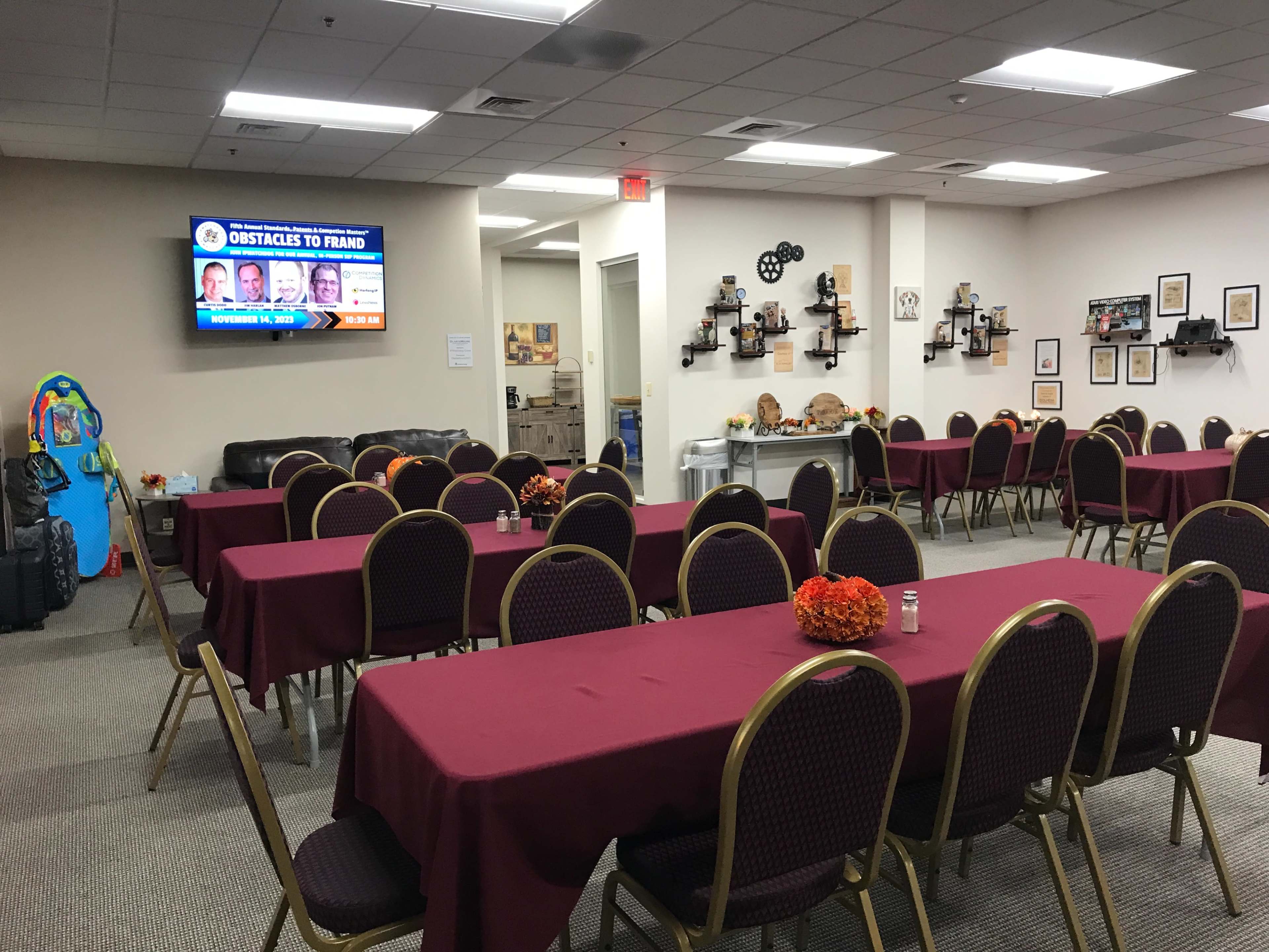 A conference room arranged with several tables covered in maroon tablecloths, with chairs set around them and a screen displaying information on a wall.