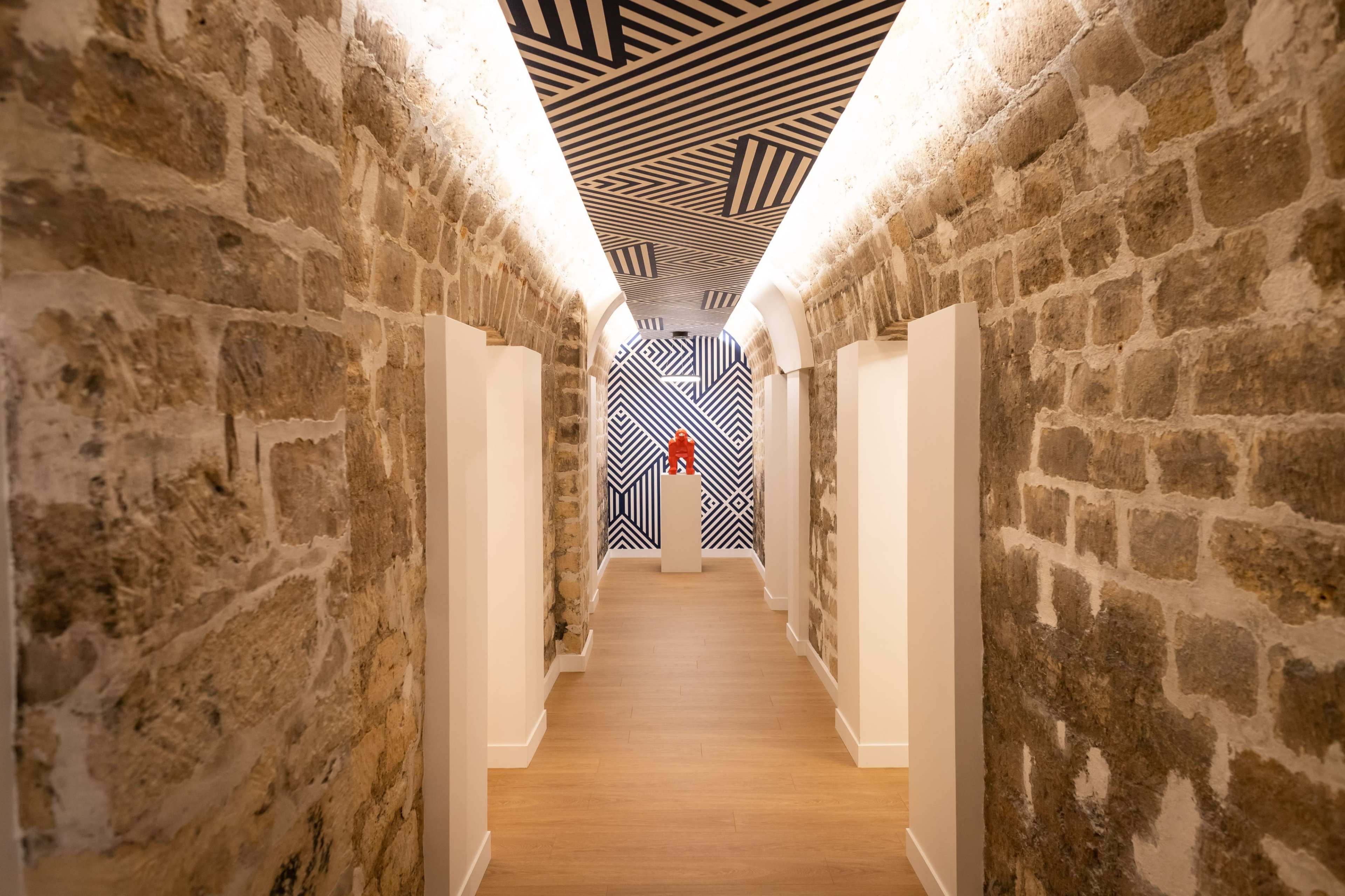 A corridor featuring exposed stone walls, a patterned ceiling, and wooden flooring, with white doors lining the sides.
