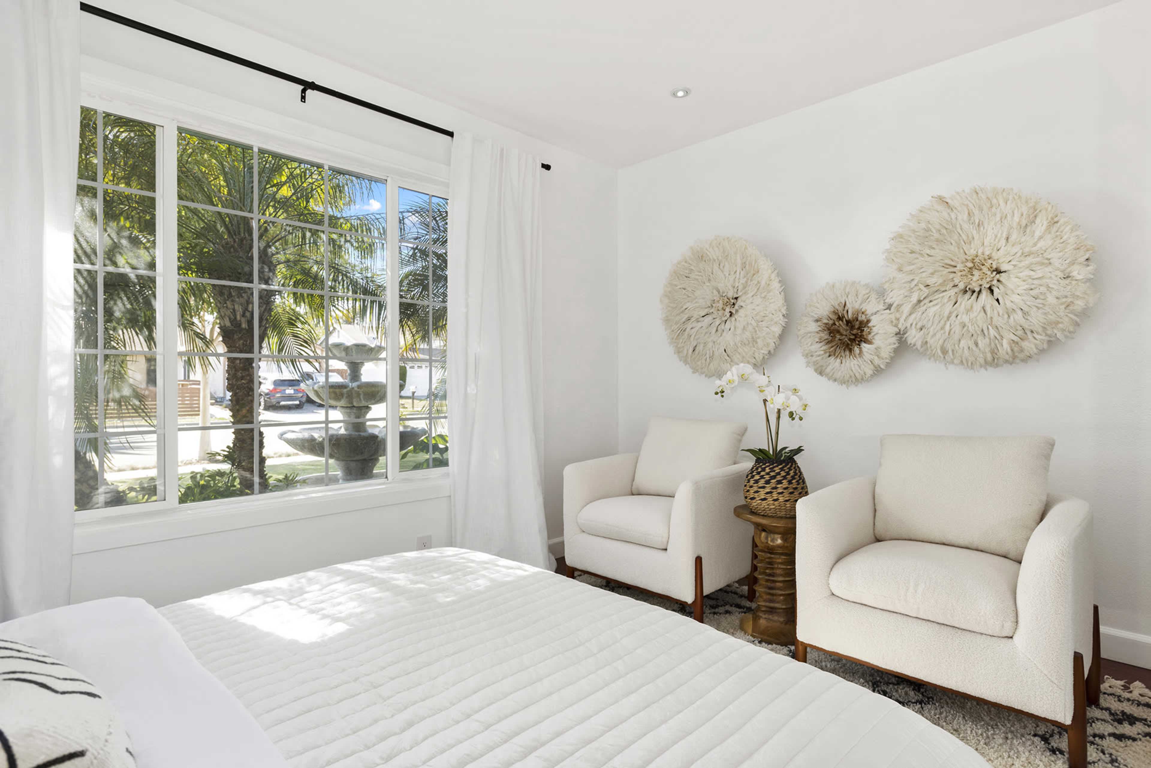 A bright bedroom features two armchairs and decorative wall art, with large windows offering a view of palm trees outside.