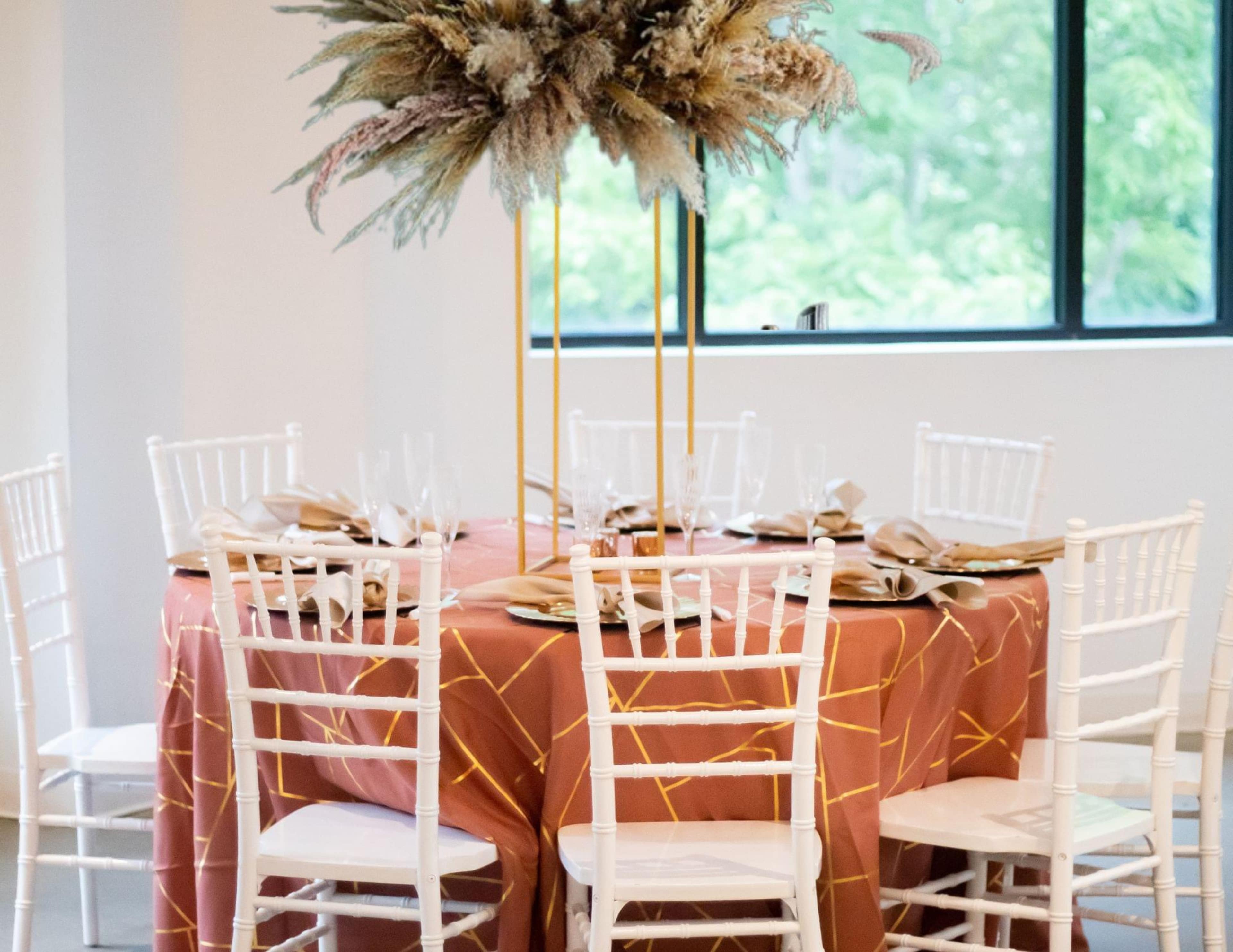 A round table with a coral tablecloth adorned with a gold geometric pattern is set for dinner, surrounded by white Chiavari chairs and featuring a floral centerpiece.