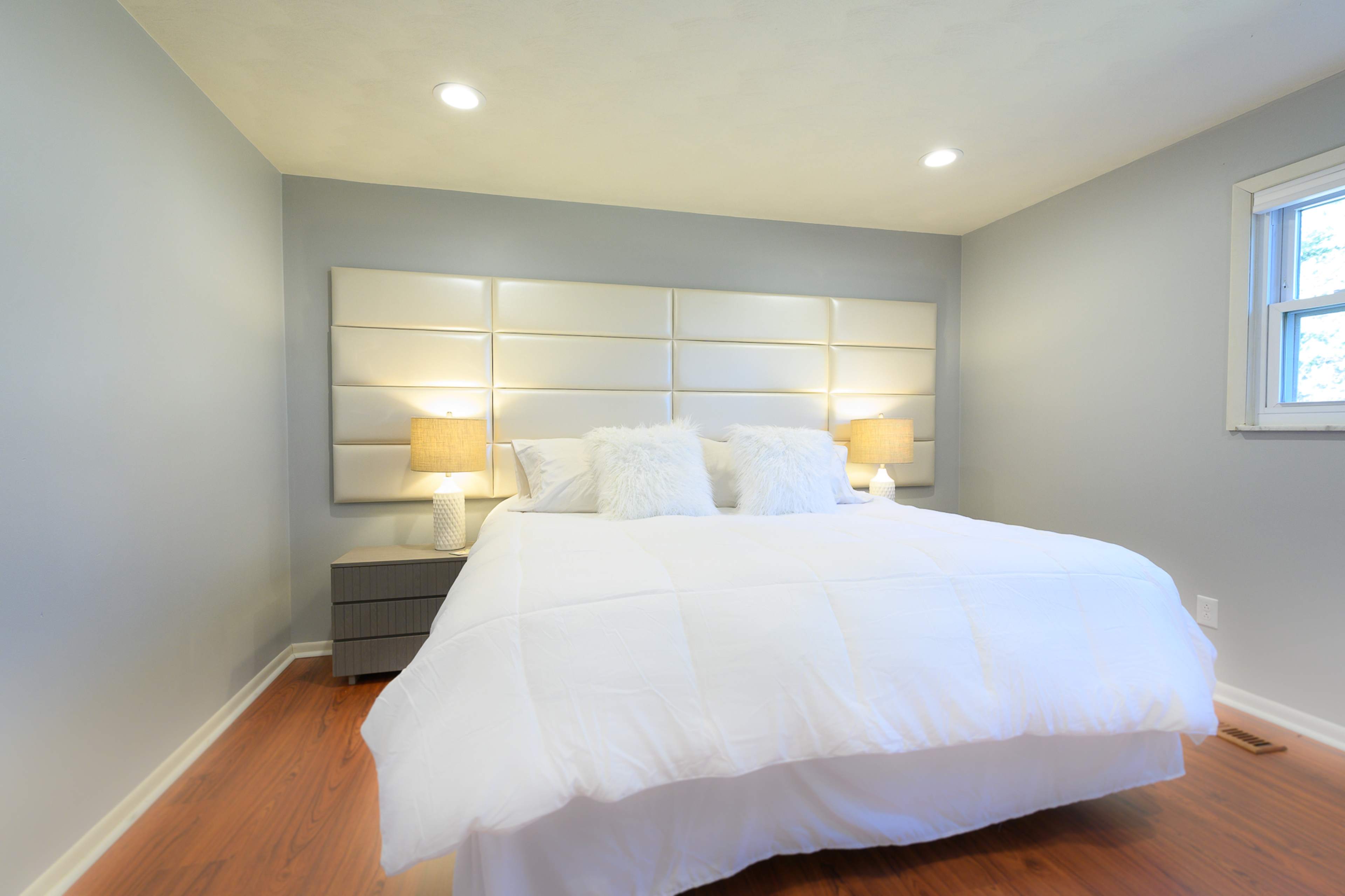 A spacious bedroom features a large bed with a white comforter, two bedside lamps, and a simple gray wall design.