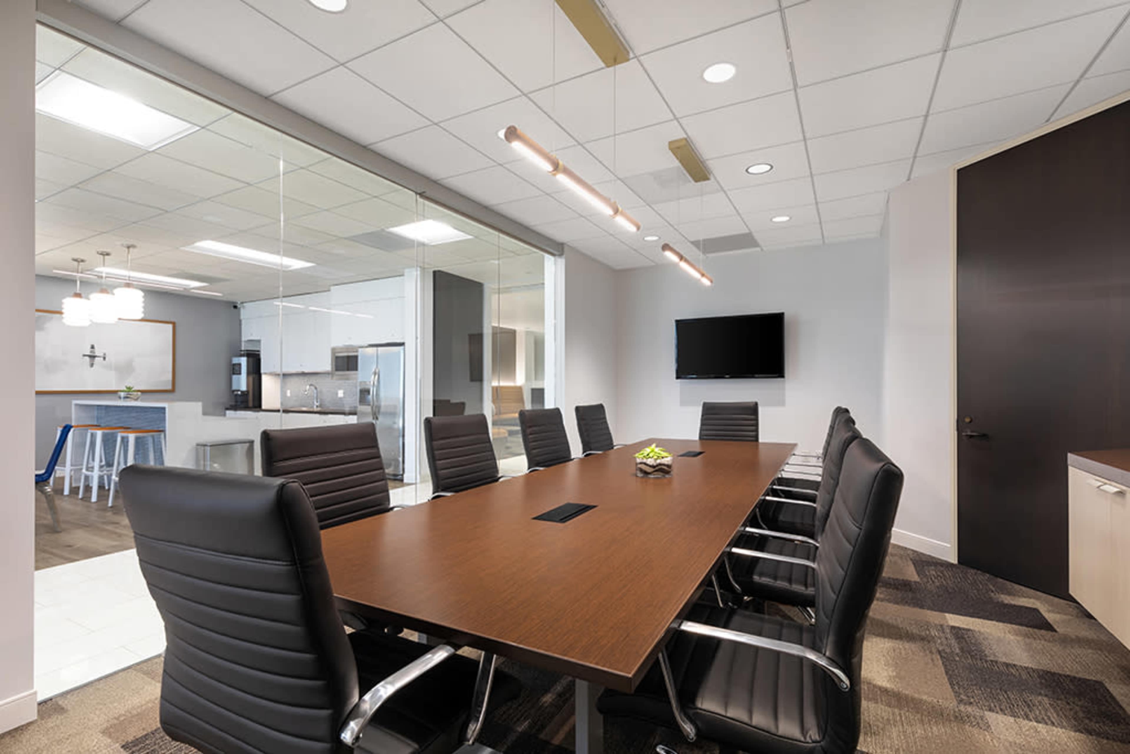 The image shows a modern conference room featuring a large wooden table surrounded by black leather chairs, with a glass wall that leads to a kitchen area visible in the background.