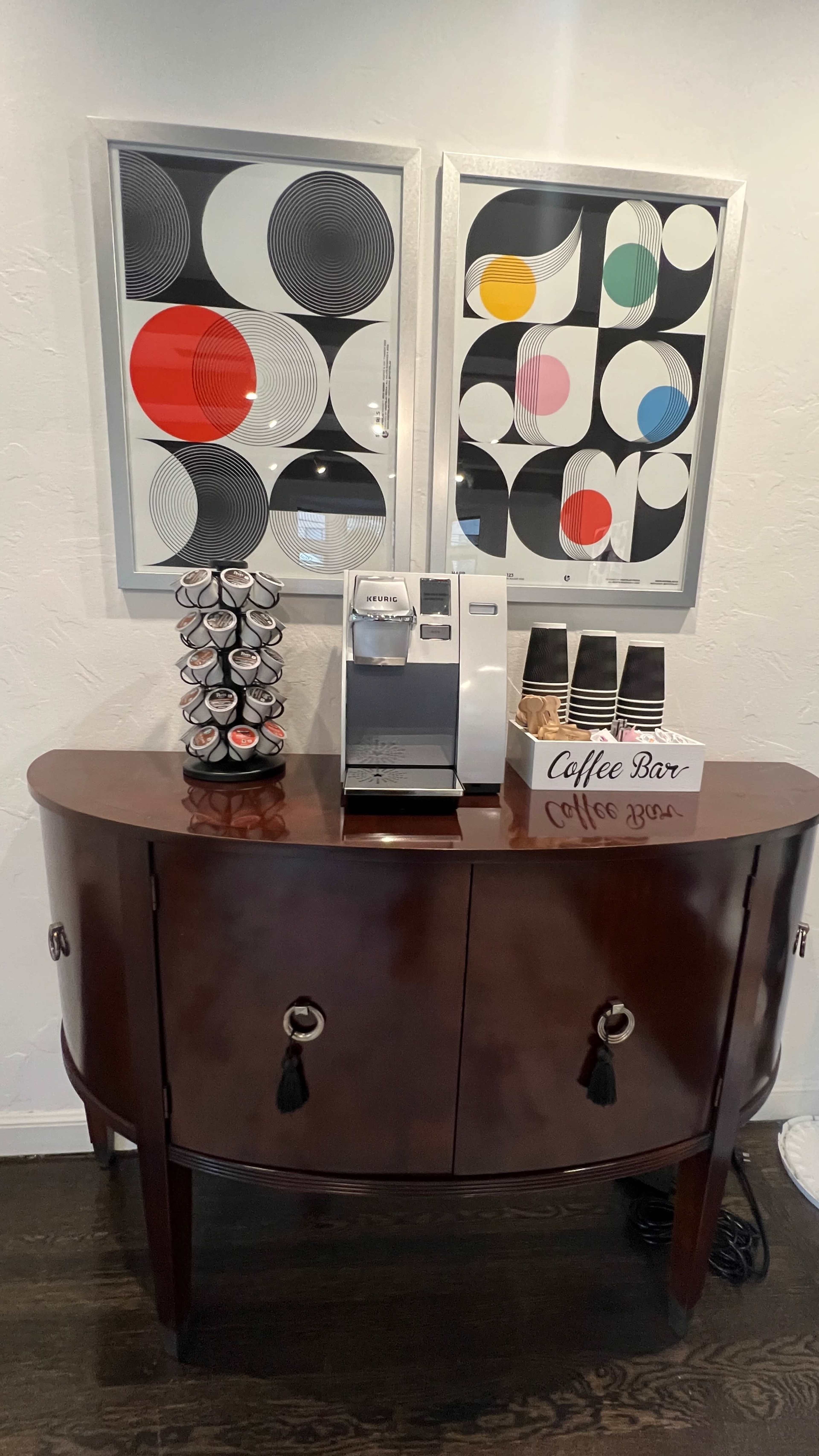 A wooden console table with a coffee machine on top, flanked by two framed geometric art pieces, and a display of coffee pods next to a labeled box reading "Coffee Bar."