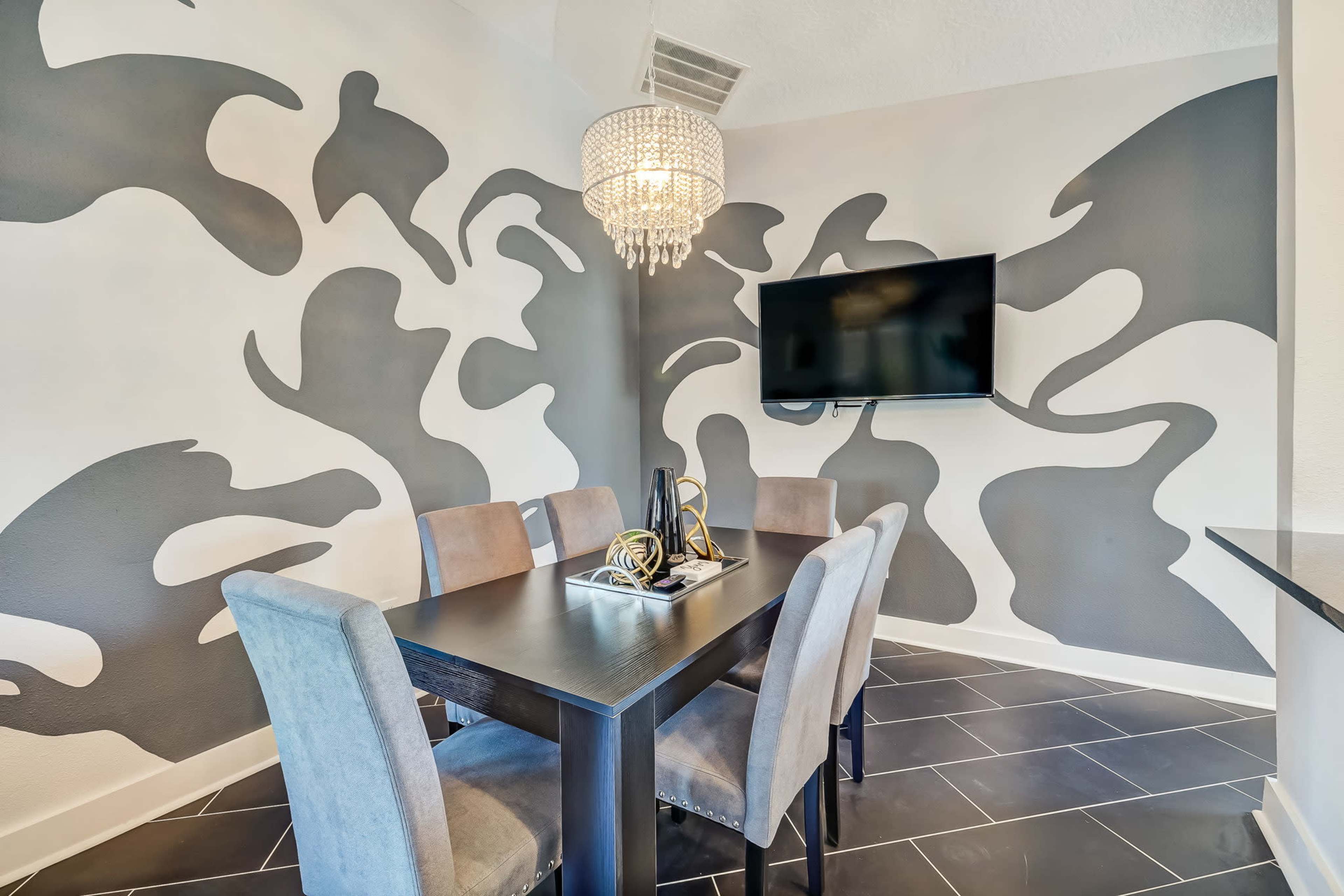 A dining area features a table with four chairs, surrounded by a patterned wall and a flat-screen TV mounted above.