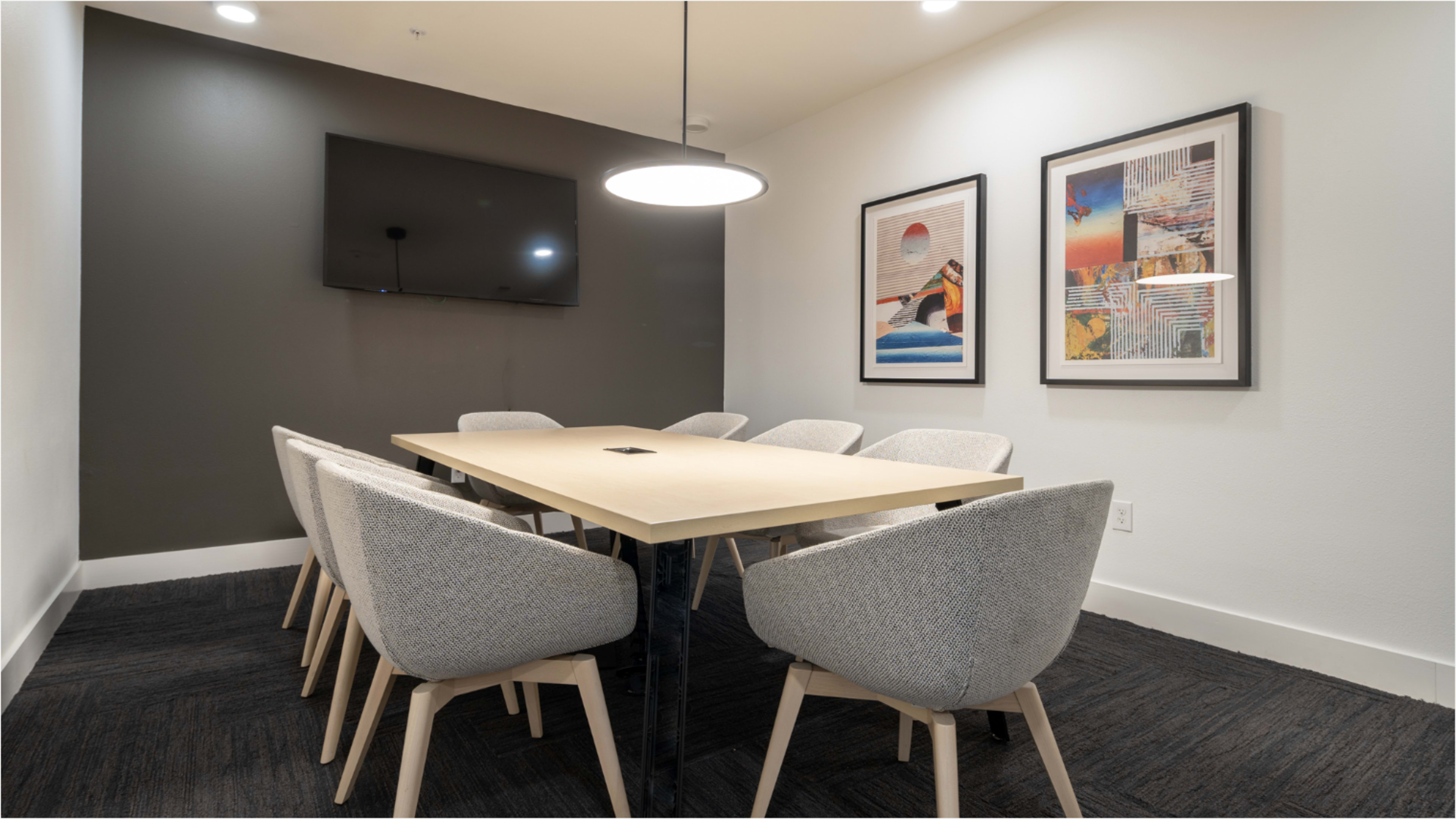 The image shows a modern conference room with a large table surrounded by eight light-colored chairs, a television mounted on the wall, and two framed artworks.