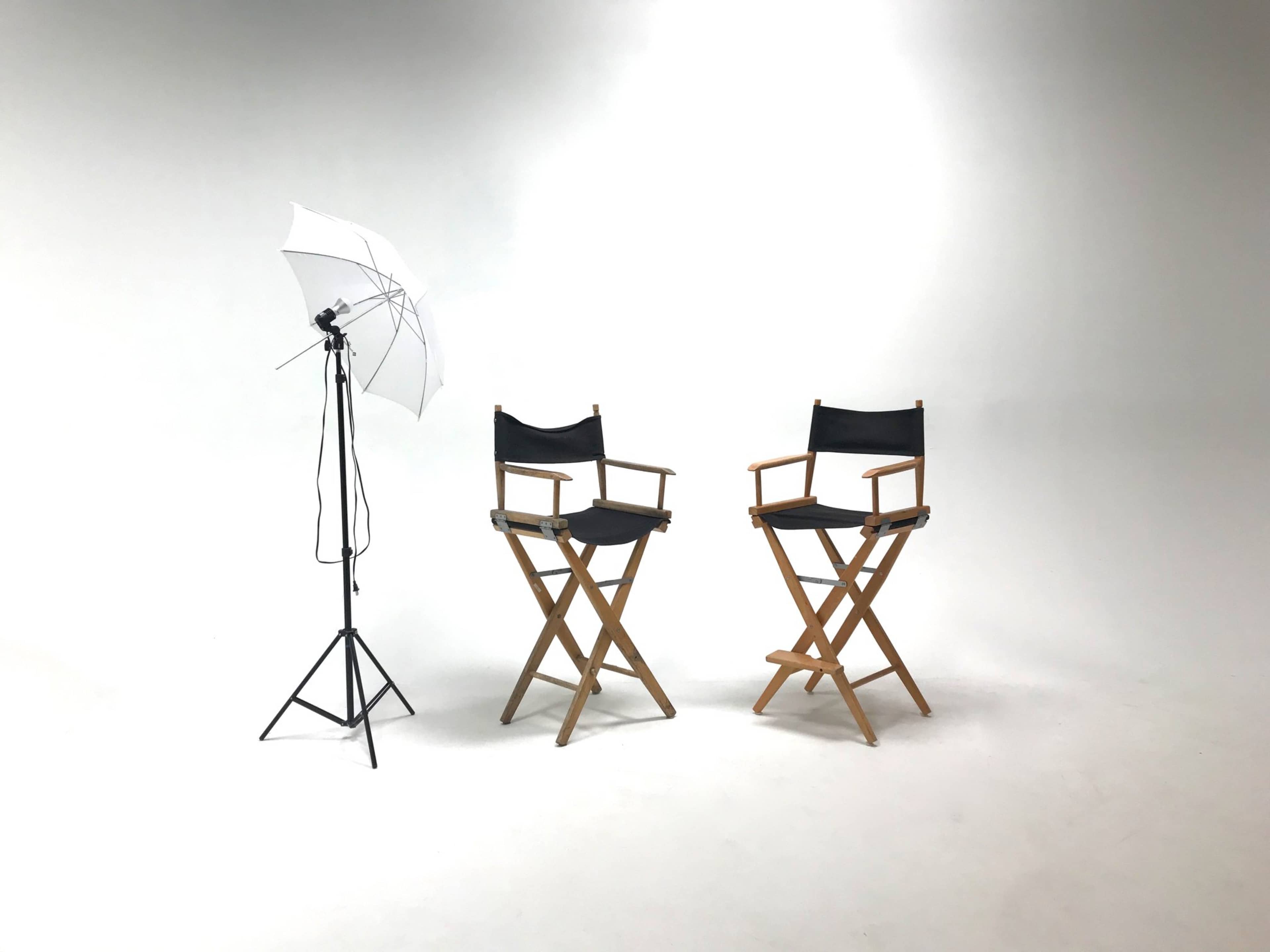 Two wooden director's chairs are placed against a backdrop of soft white light, next to a photography umbrella on a stand.