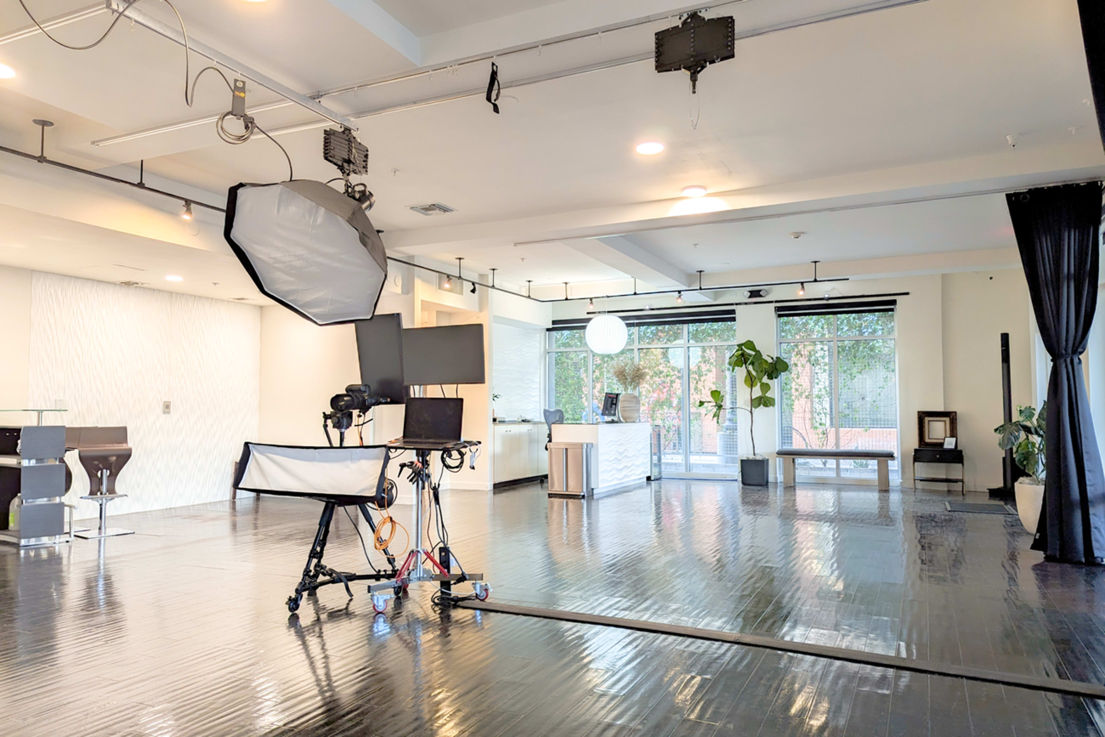 A spacious room is set up with photography equipment, including a softbox and monitors, alongside large windows featuring plants and natural light.