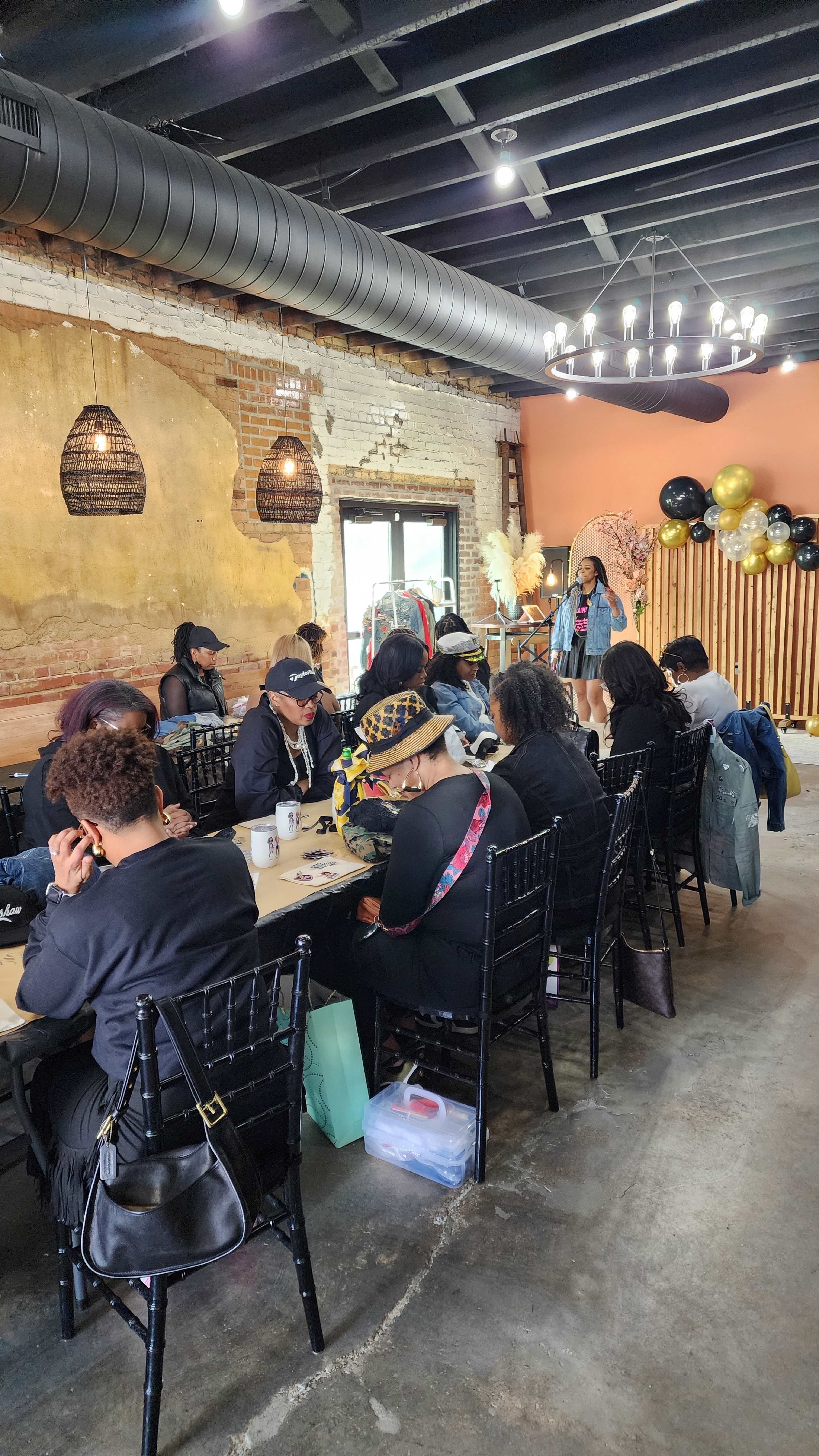 A group of people is seated at a long table in a cafe, engaged in conversation and activities, with black and gold decorations visible in the background.
